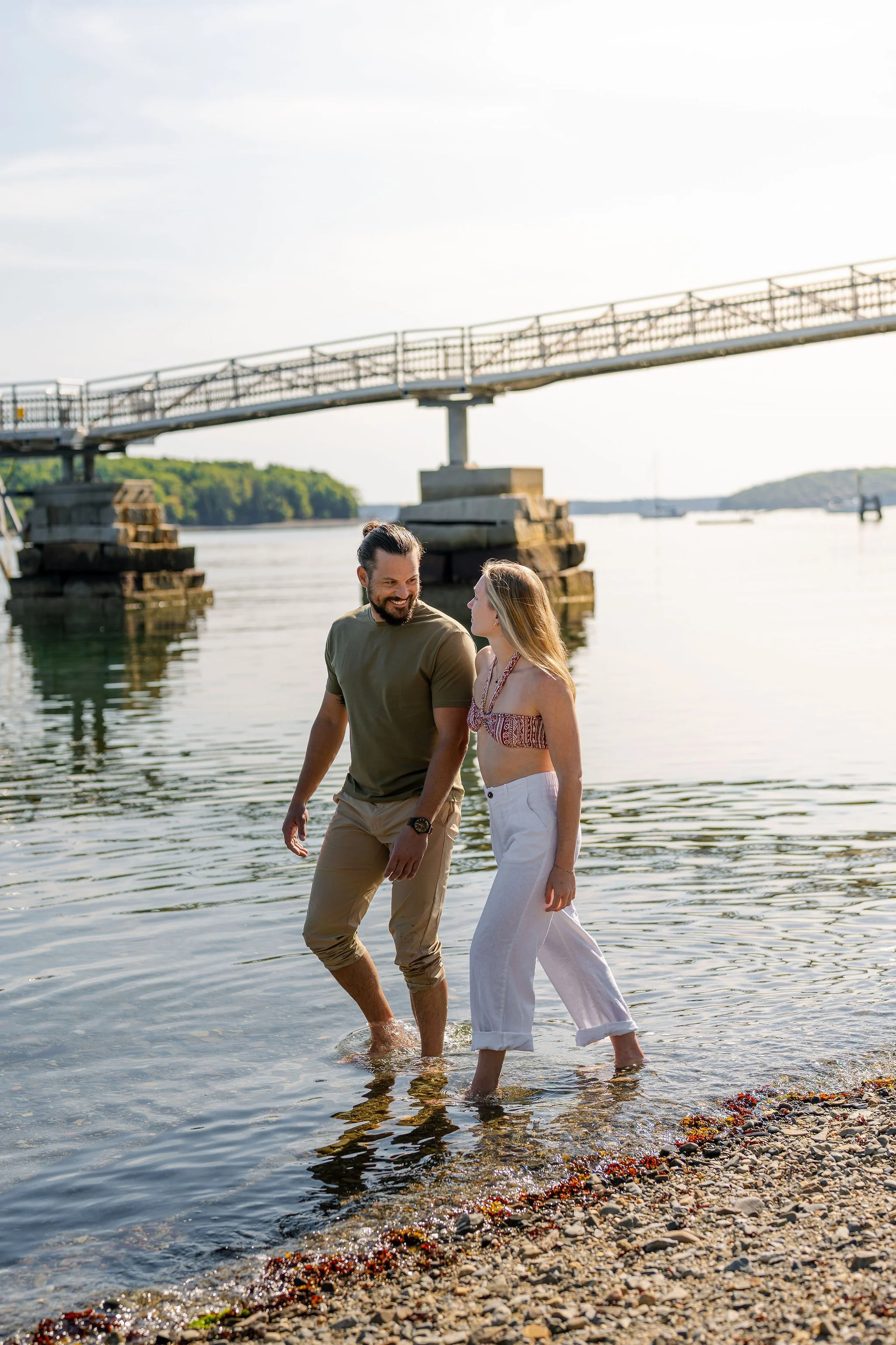 A man and woman are standing in shallow water at a beach or lakeside, smiling at each other. The man is wearing a green t-shirt and beige pants rolled up to his knees, while the woman is in a patterned crop top and white pants. There is a bridge in t