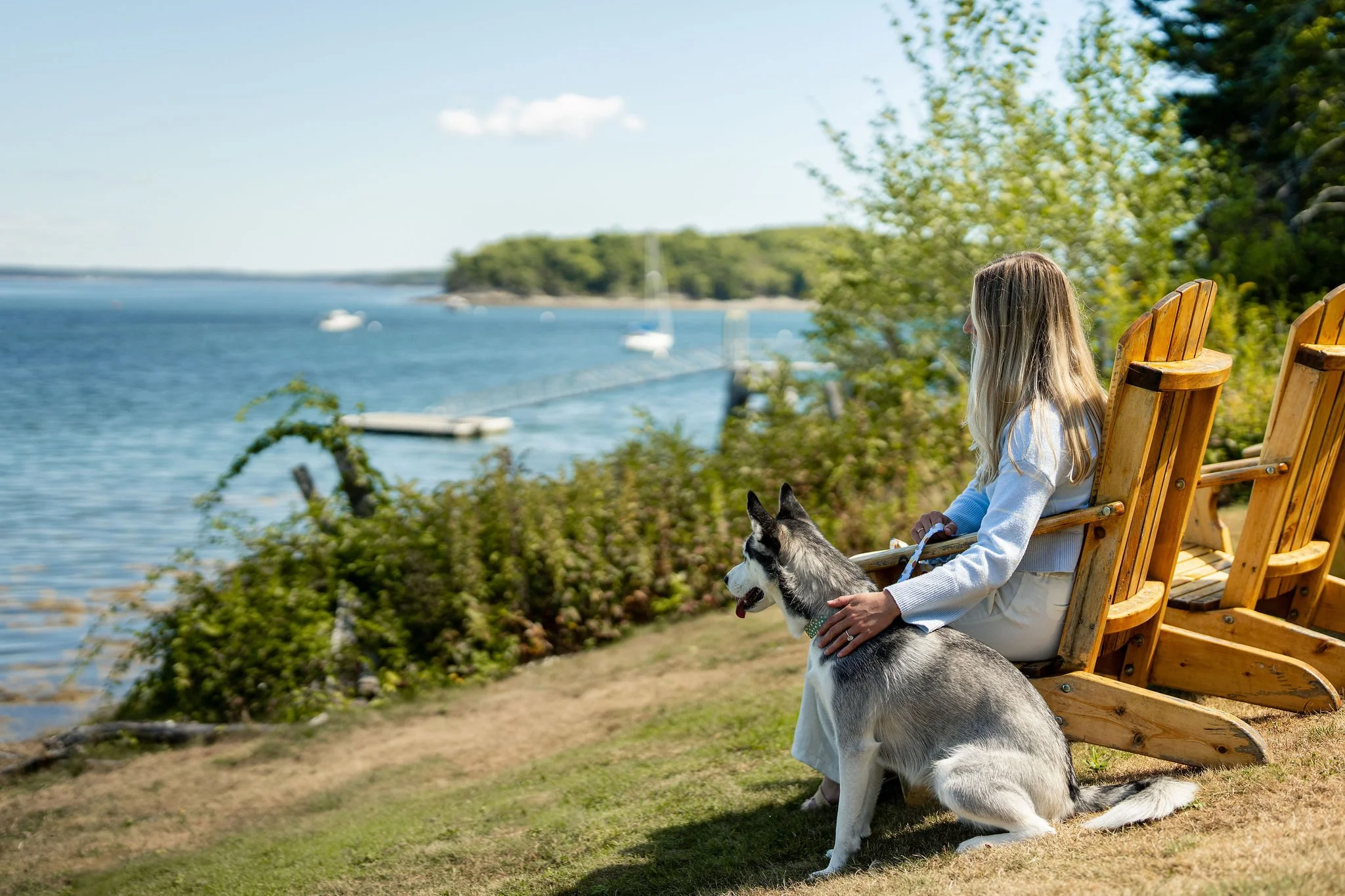 A woman with long blonde hair sitting on a wooden chair near a body of water, with a Siberian Husky sitting beside her. The setting is outdoors, on a grassy area with trees and boats visible in the water in the background.
