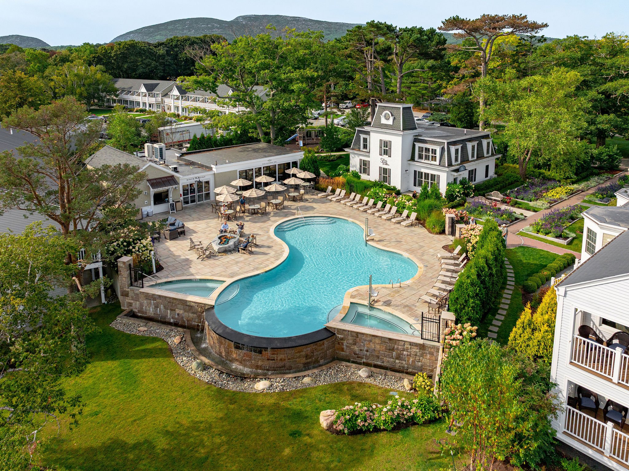 Aerial view of a resort swimming pool with lounge chairs, umbrellas, a hot tub, surrounded by lush greenery, small buildings, and a white mansion in the background.
