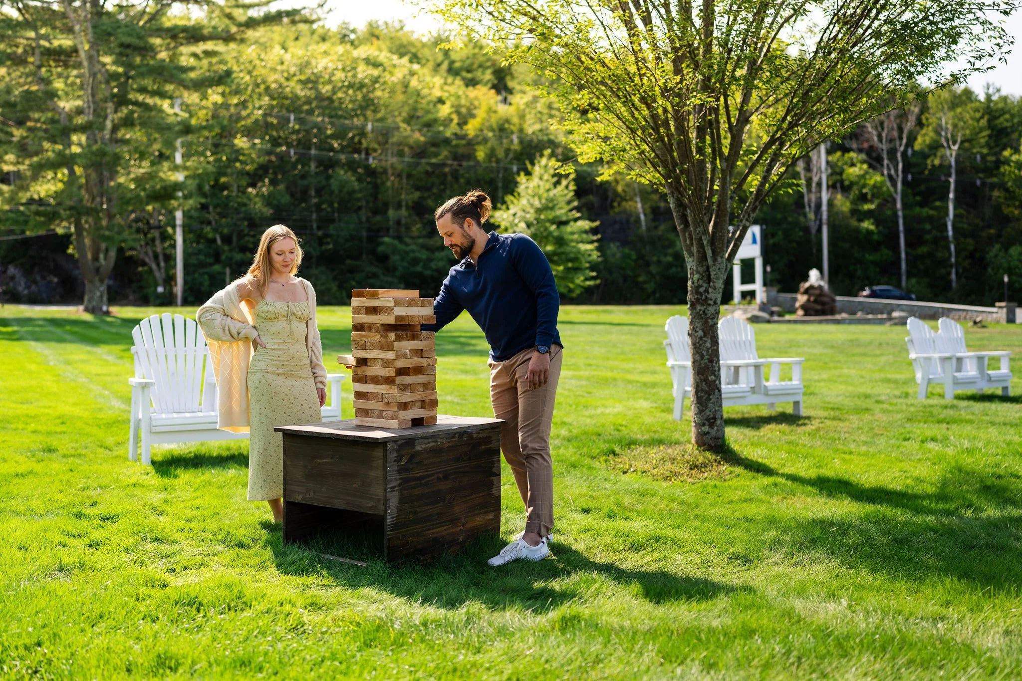 Two people playing giant Jenga outdoors on a sunny day with green grass and trees around, and white benches in the background.