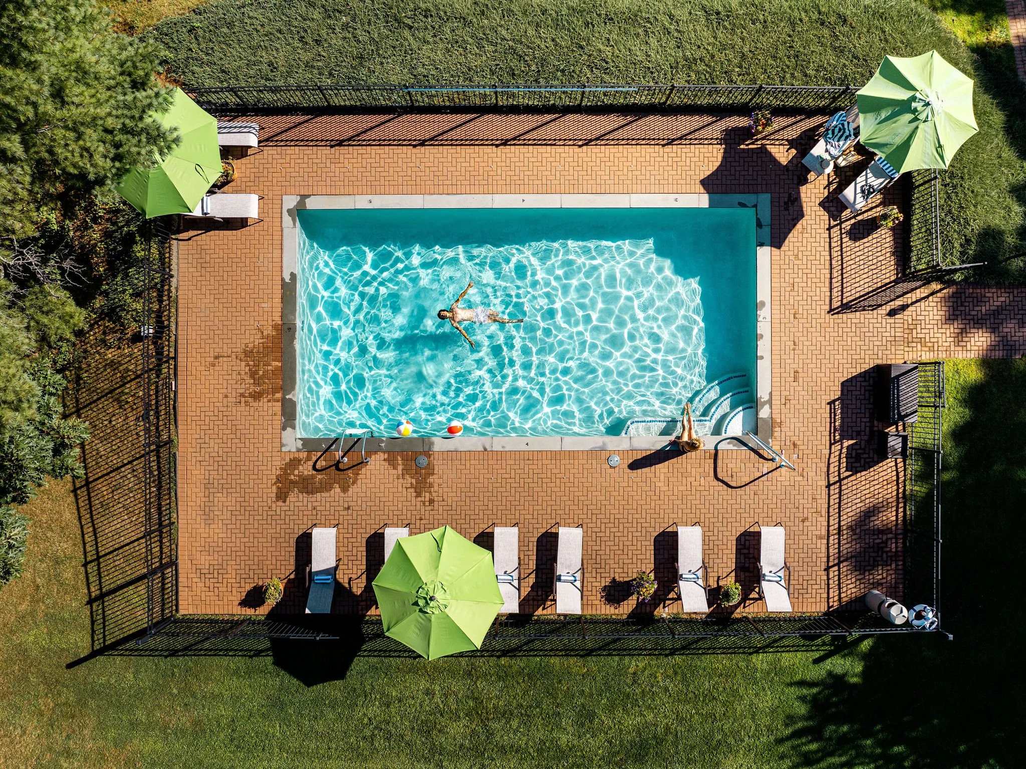An aerial view of a small swimming pool with two people swimming and relaxing. The pool is surrounded by a brick patio with several lounge chairs, green umbrellas, and potted plants. The area is enclosed with a black metal fence, and there are trees 
