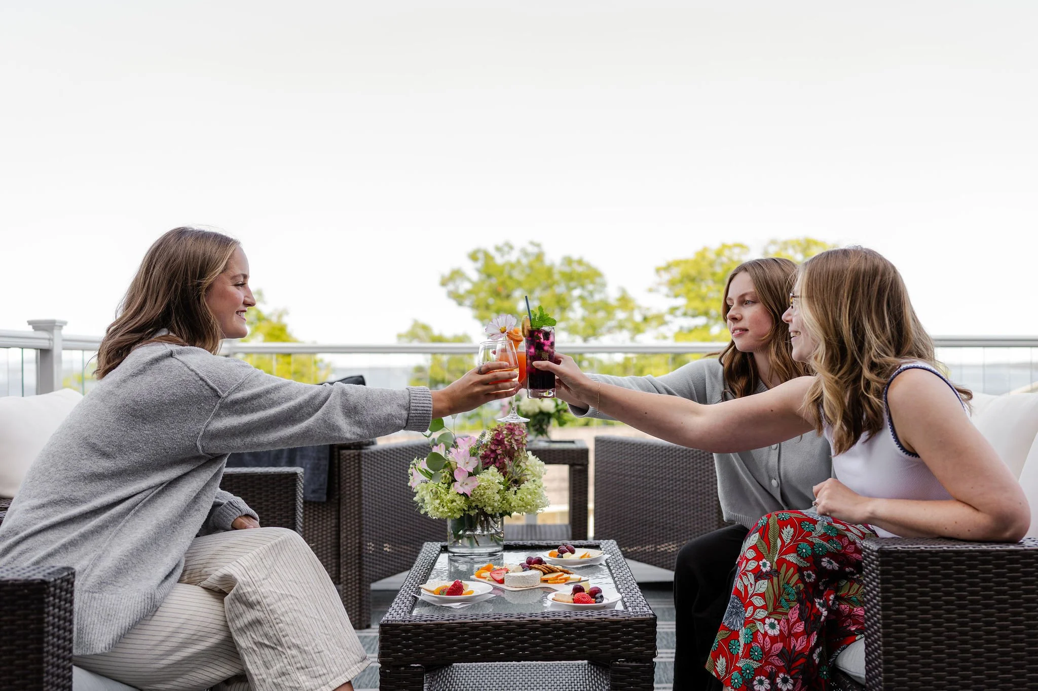 Three women having drinks and toasts, sitting on a patio with a table of snacks and flowers, outdoors on a sunny day.