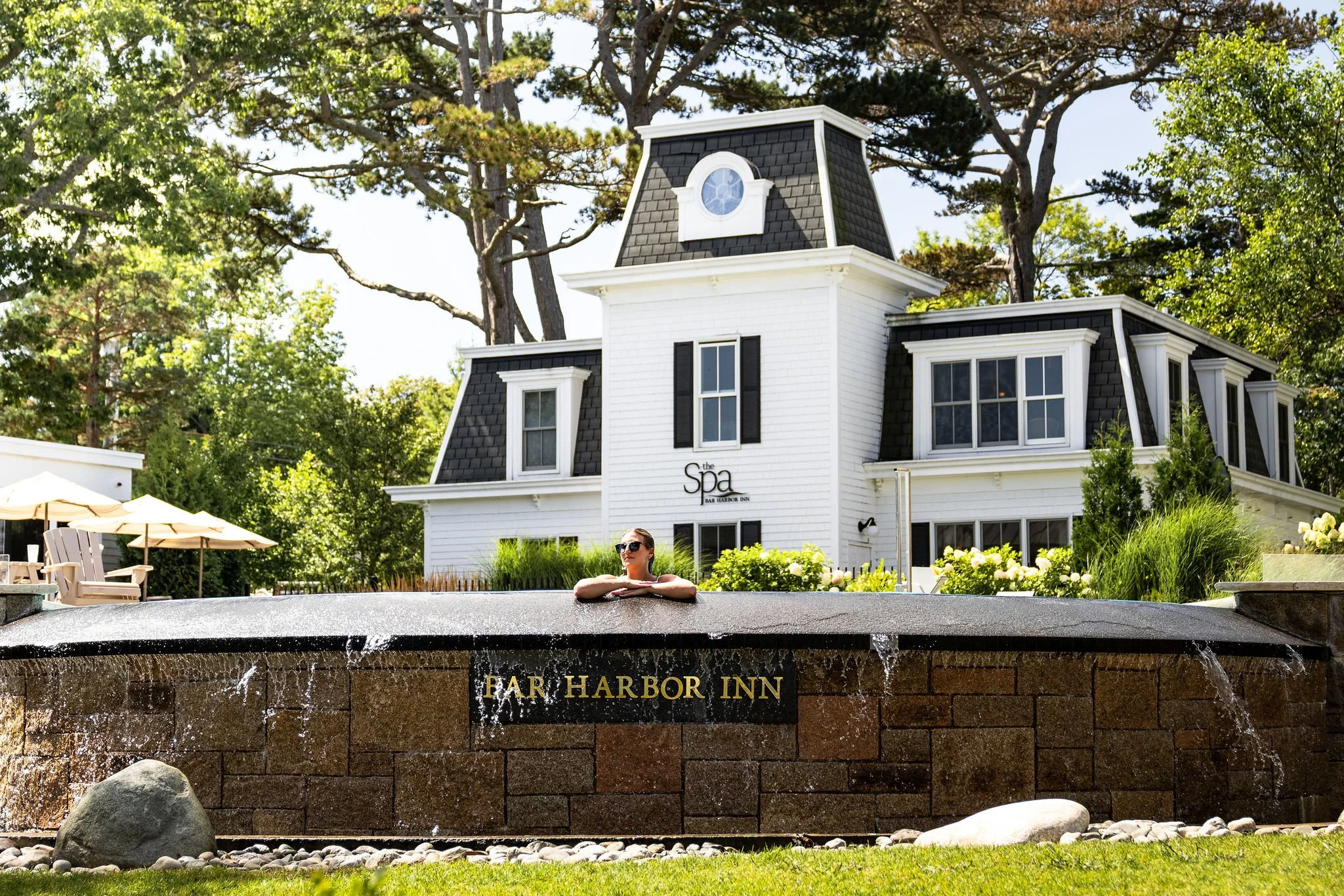 A woman wearing sunglasses relaxing at the edge of an outdoor hot tub with the sign 'Far Harbor Inn' on a brick wall and a white building with black accents and a turret in the background, surrounded by trees and greenery.
