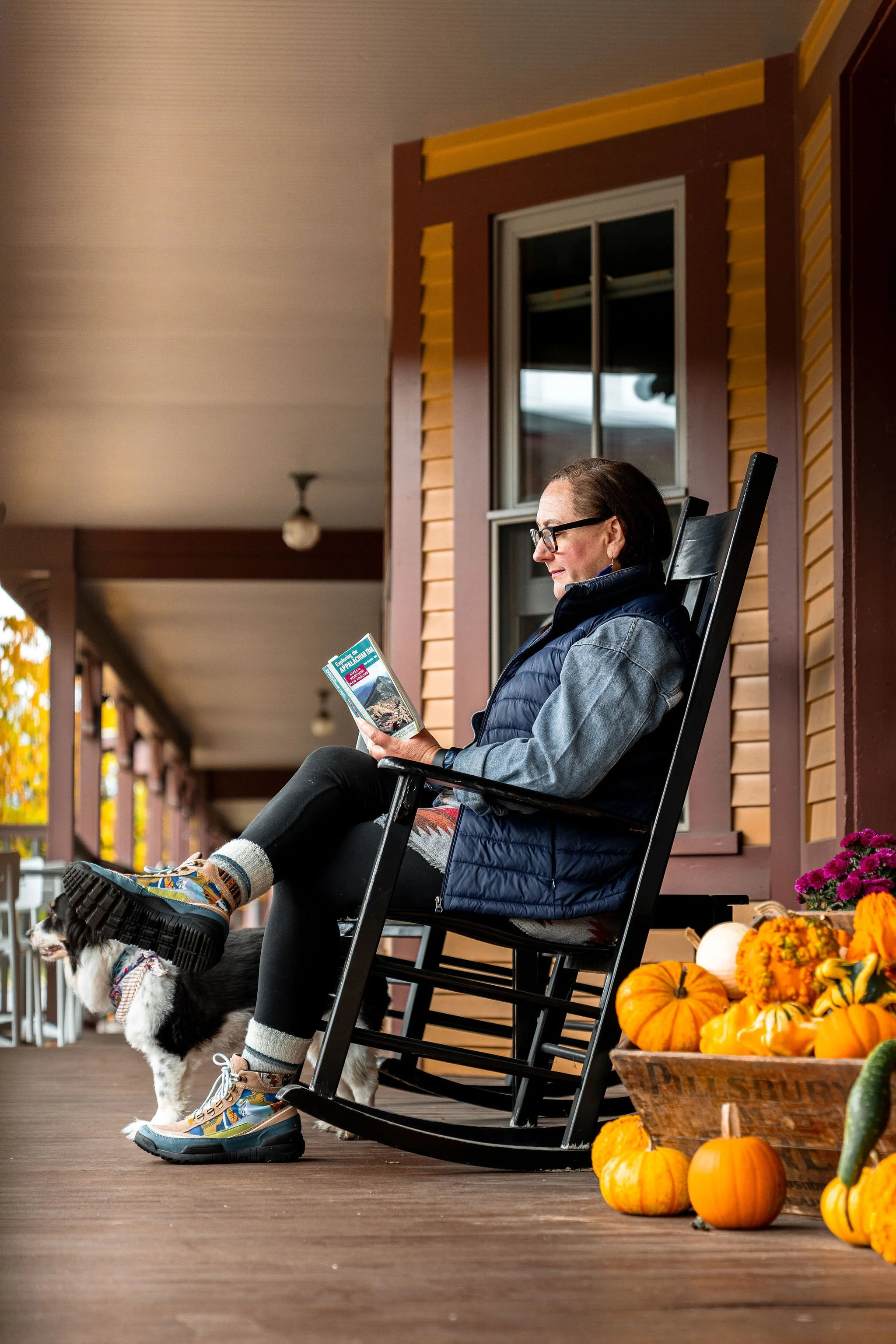 A woman sitting on a rocking chair on a porch, reading a book with a dog nearby, and pumpkins and flowers in a wooden crate beside her.