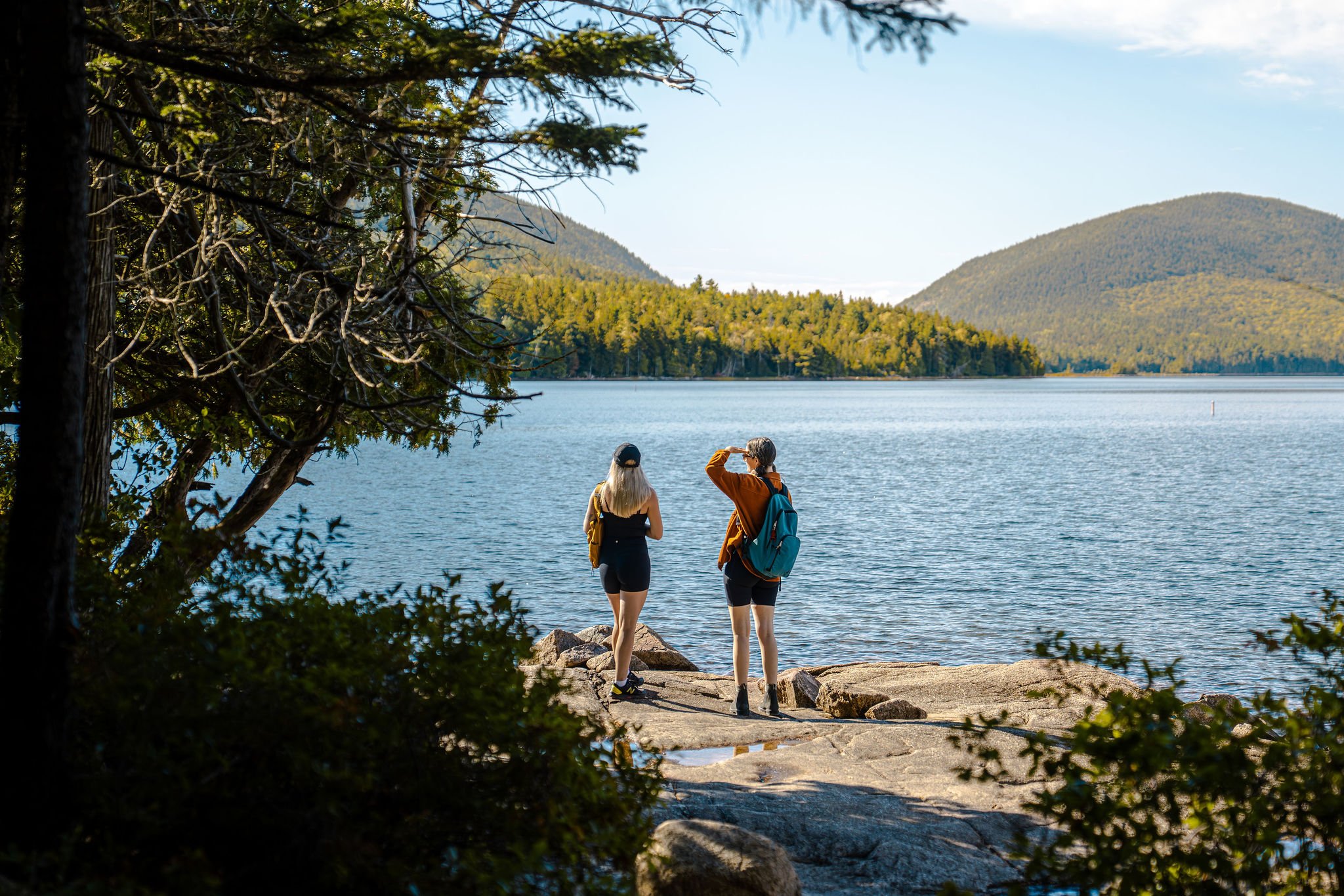 Two women with backpacks standing on rocks by a lake, surrounded by trees, with mountains in the background, enjoying the view.