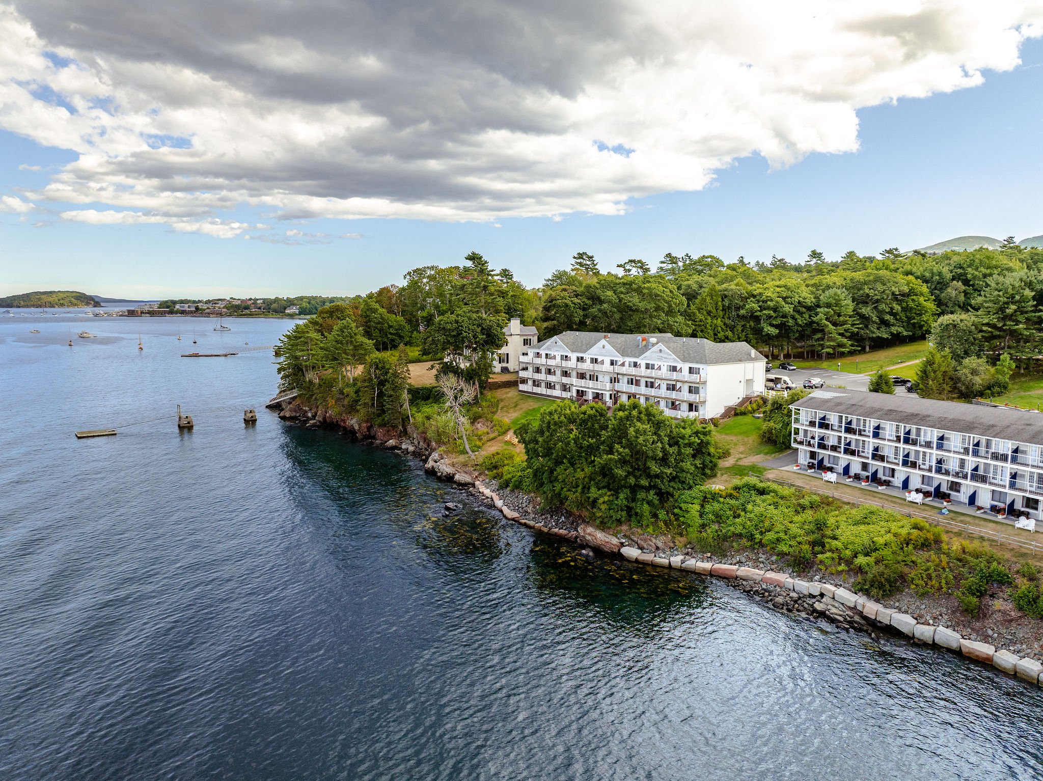 Aerial view of a waterfront with a white apartment complex, green trees, rocky shoreline, and boats anchored in the water under a partly cloudy sky.
