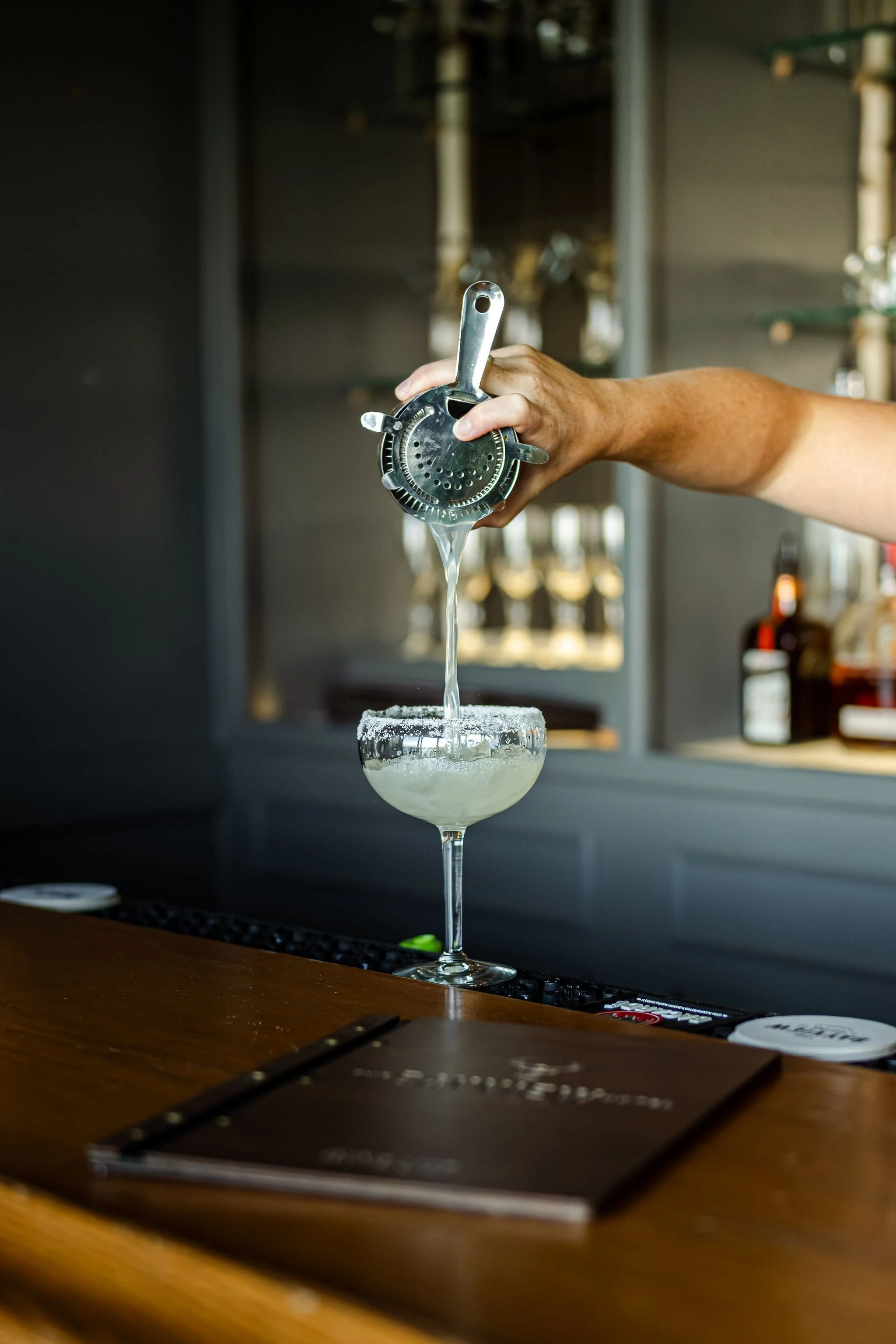 Bartender pouring a cocktail through a cocktail strainer into a salted rim glass, with a menu on the wooden bar in the foreground.