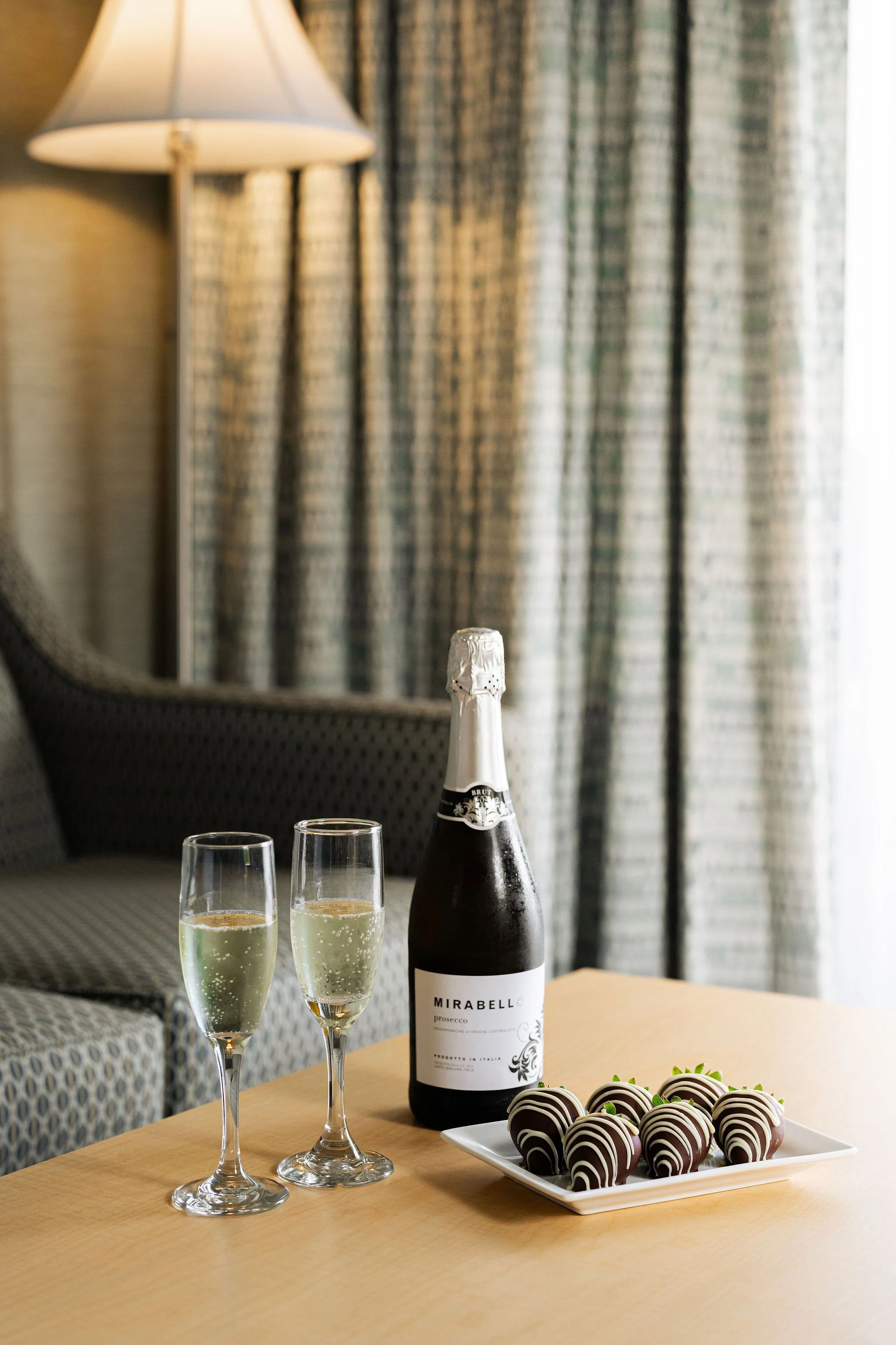 A bottle of Mirabello prosecco, two glasses of bubbly, and a plate of chocolate-covered strawberries on a wooden table, with a sofa, patterned curtains, and a lamp in the background.