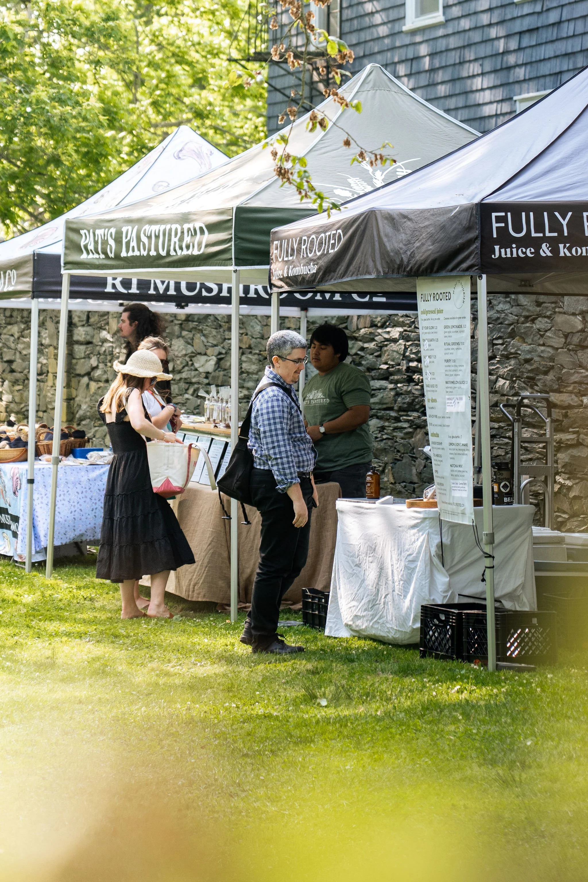 People browsing at outdoor market stalls with tents that have signs reading 'PATS PASTURED', 'FULLY ROOTED', and 'JUICE & KOMBUCHA', set against a stone wall and green trees.