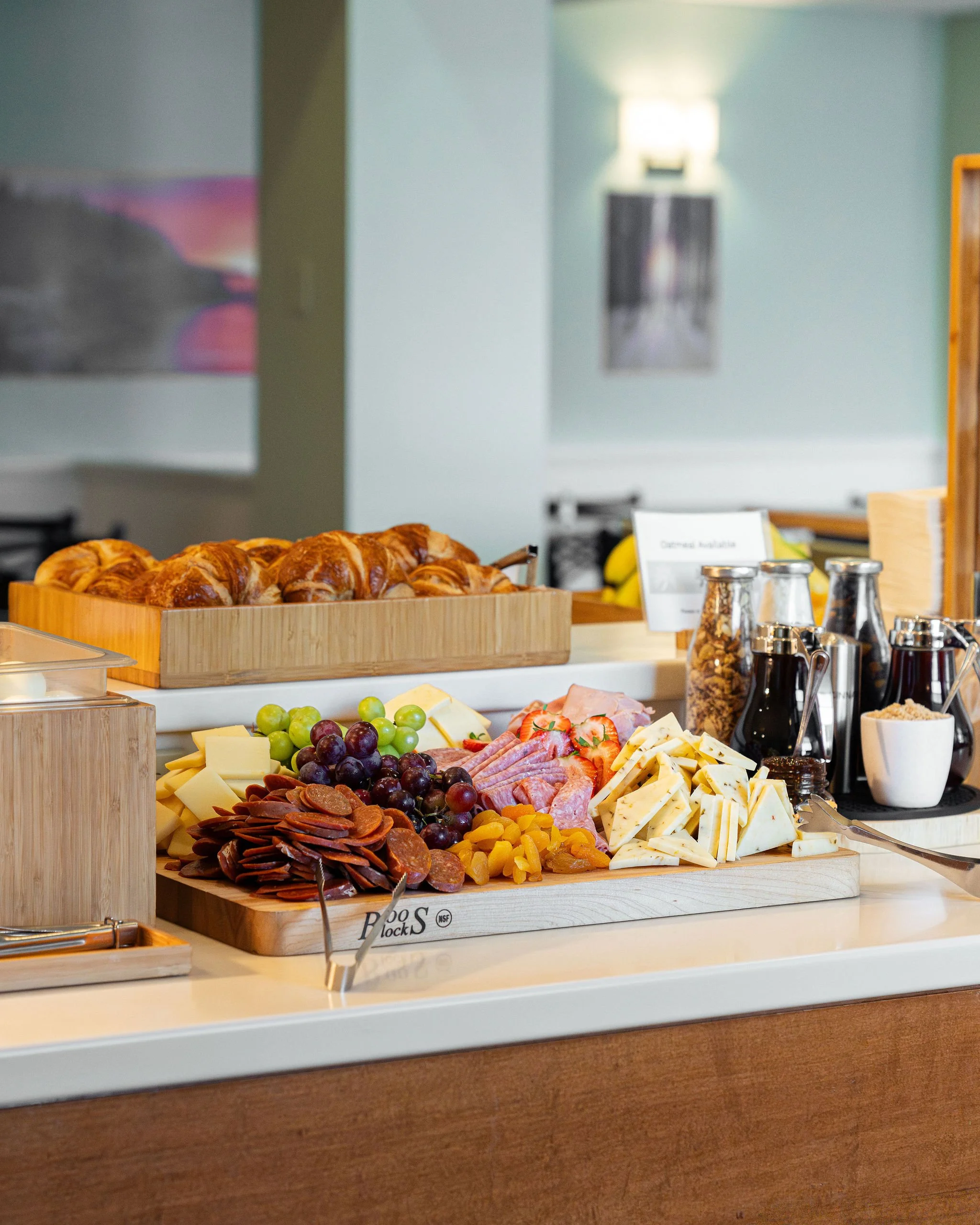 A breakfast buffet with croissants, a cheese platter with grapes, sliced meats, and jams, alongside coffee urns and cereal jars, on a white countertop in a dining area.