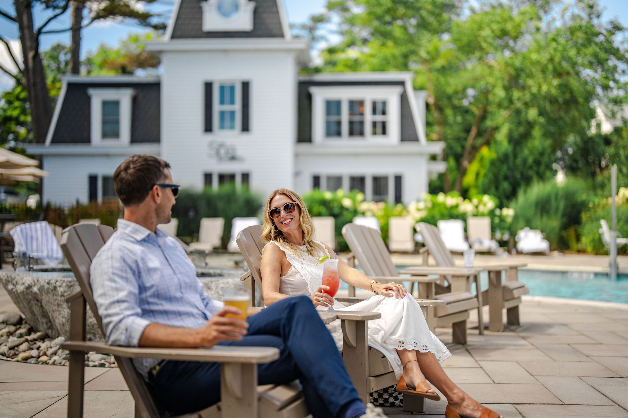 A man and woman relaxing by a pool on lounge chairs, enjoying drinks, with a white house, trees, and a pool in the background.