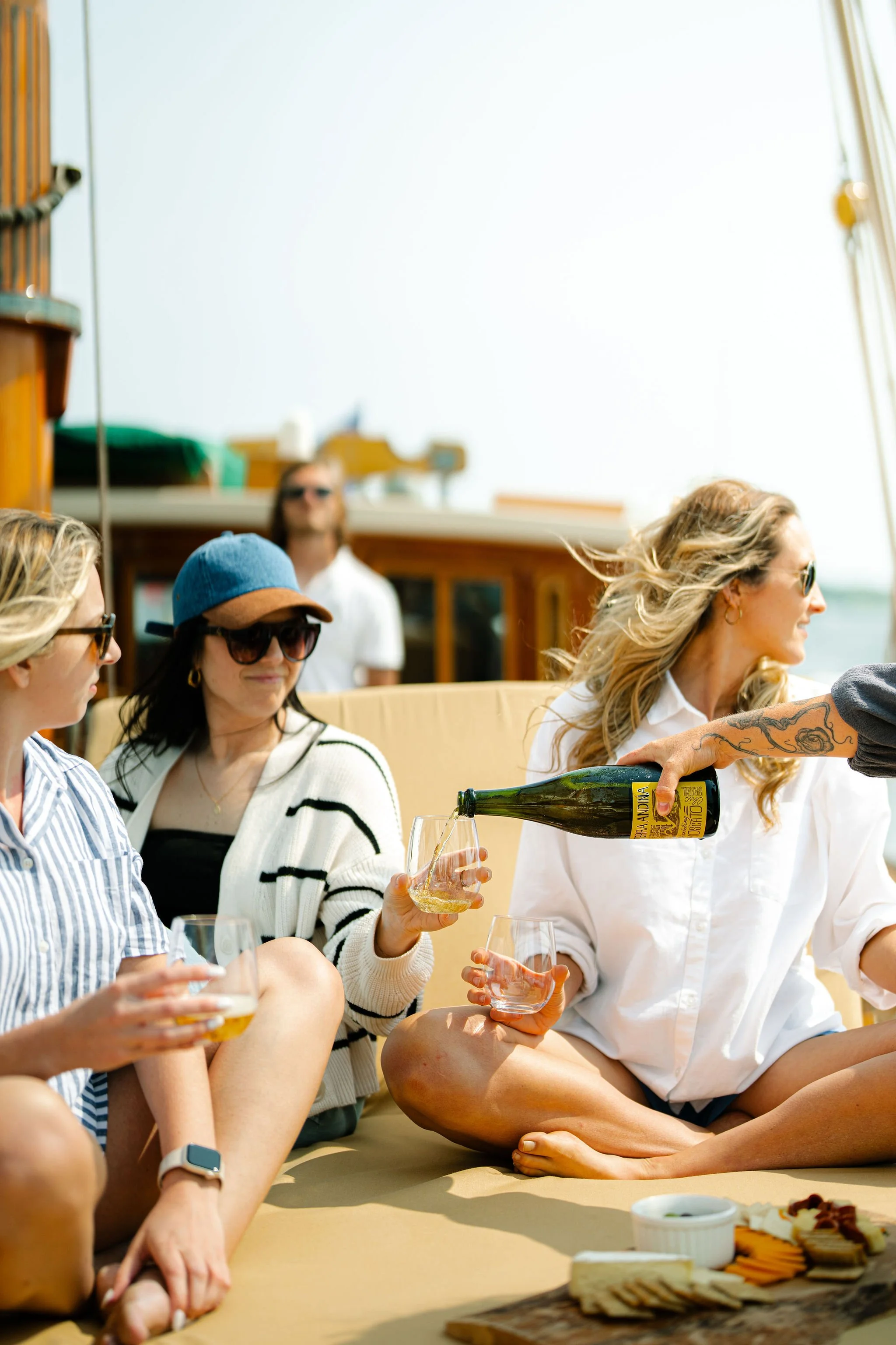 People relaxing on a boat with snacks and drinks, pouring champagne, with a woman in sunglasses and a striped shirt on the left, a woman with a blue cap and sunglasses in the middle, and a woman with long hair in white on the right.