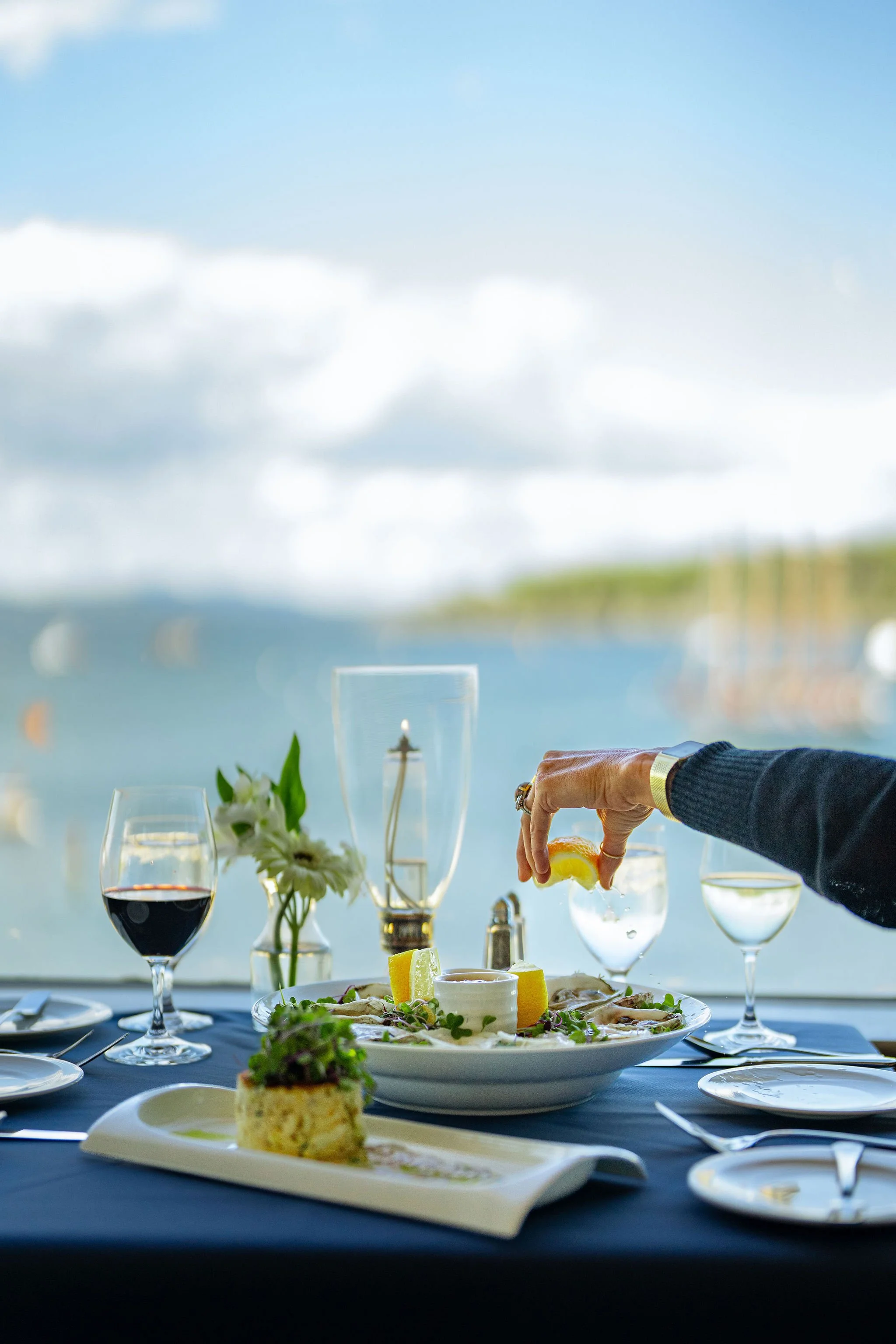 A dining table set with wine glasses, a floral centerpiece, and a plate of food, with a scenic water view and cloudy sky in the background.