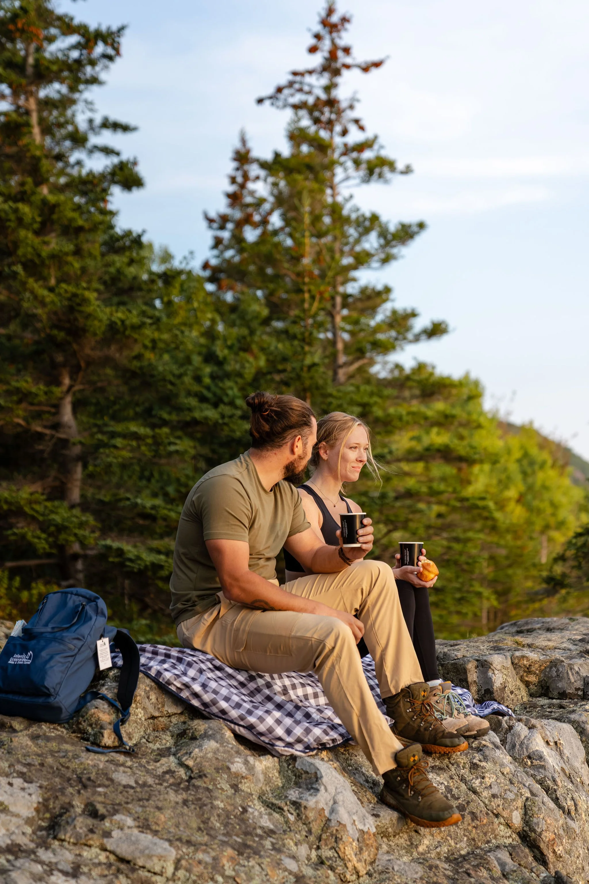 A man and woman sitting on rocks in a forest, having coffee and a snack, with a backpack nearby, during sunset.