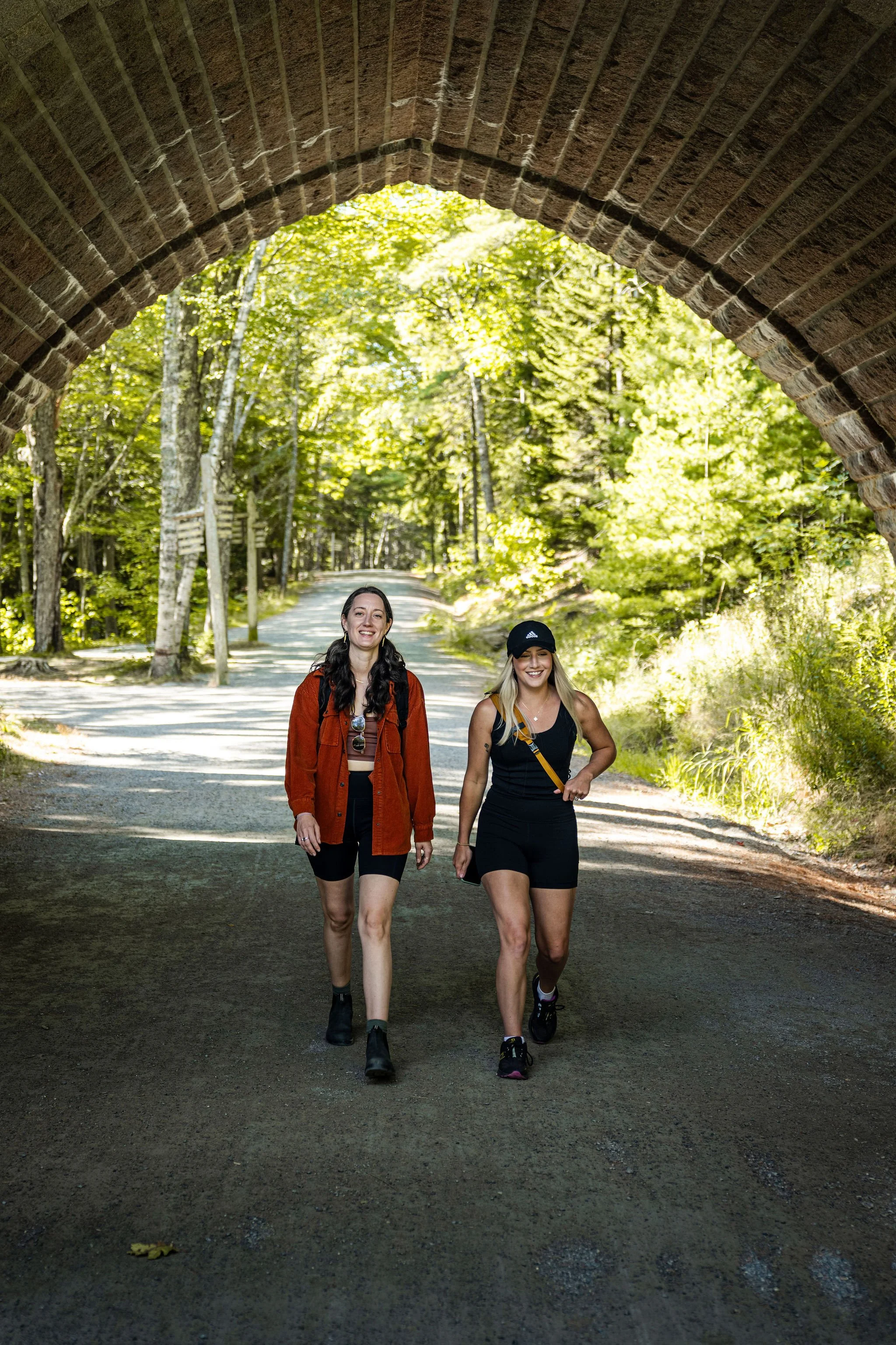Two women walk on a dirt path under a brick tunnel, surrounded by lush green trees in a forest.