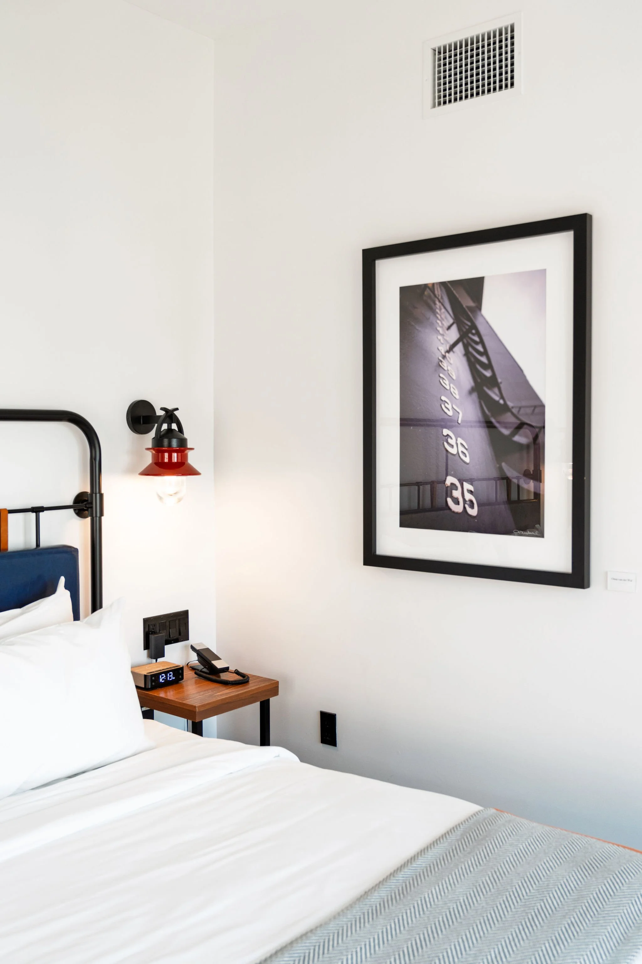 A hotel room featuring a bed with white bedding, a bedside table with a digital clock, phone, and light switch, a wall-mounted lamp, a framed photograph of a speedometer, a white wall, and a ceiling vent.