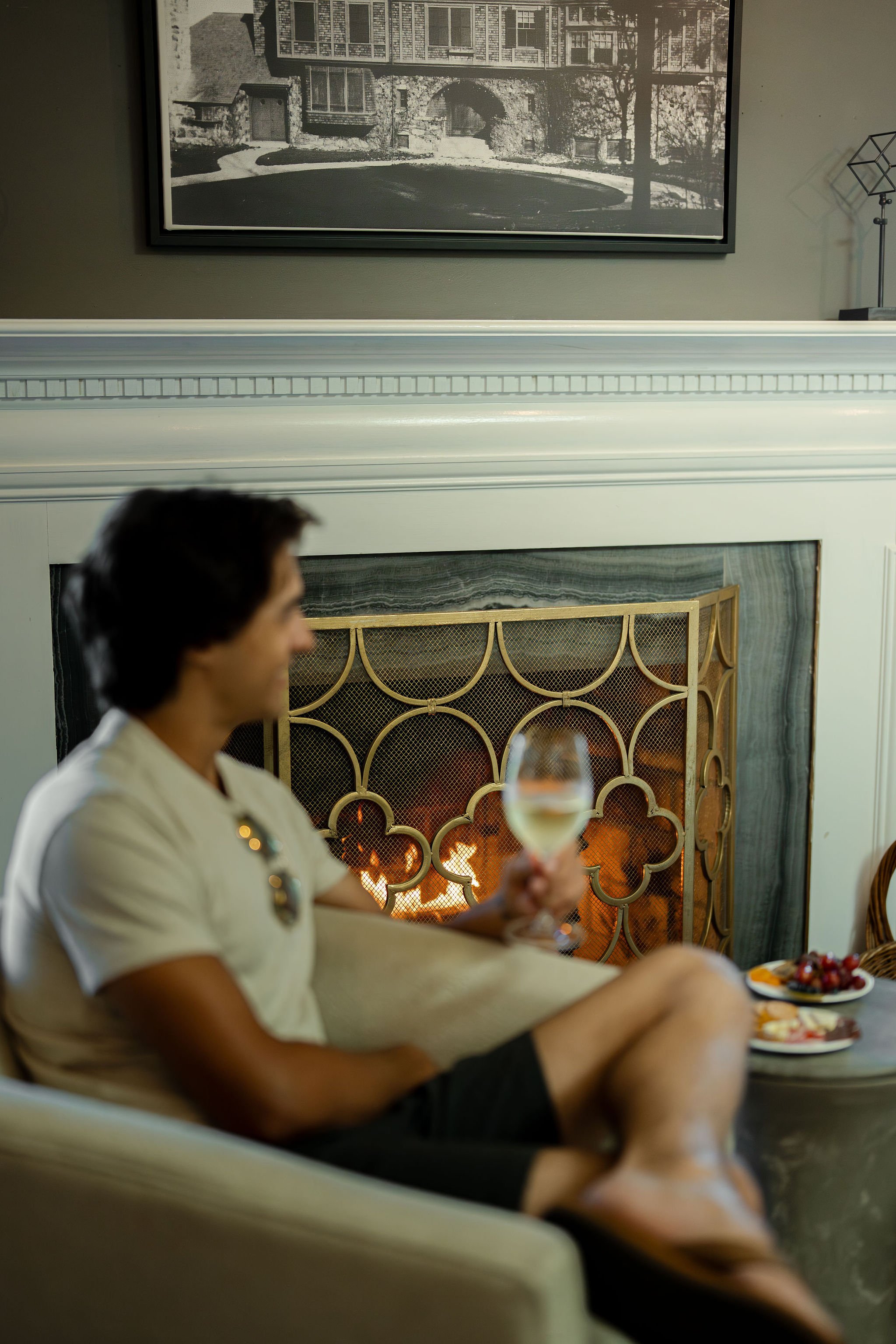 A man sitting on a beige chair by a fireplace, holding a glass of white wine, with a plate of cheese and grapes on a table nearby, in a cozy living room with a framed black and white photo on the wall.
