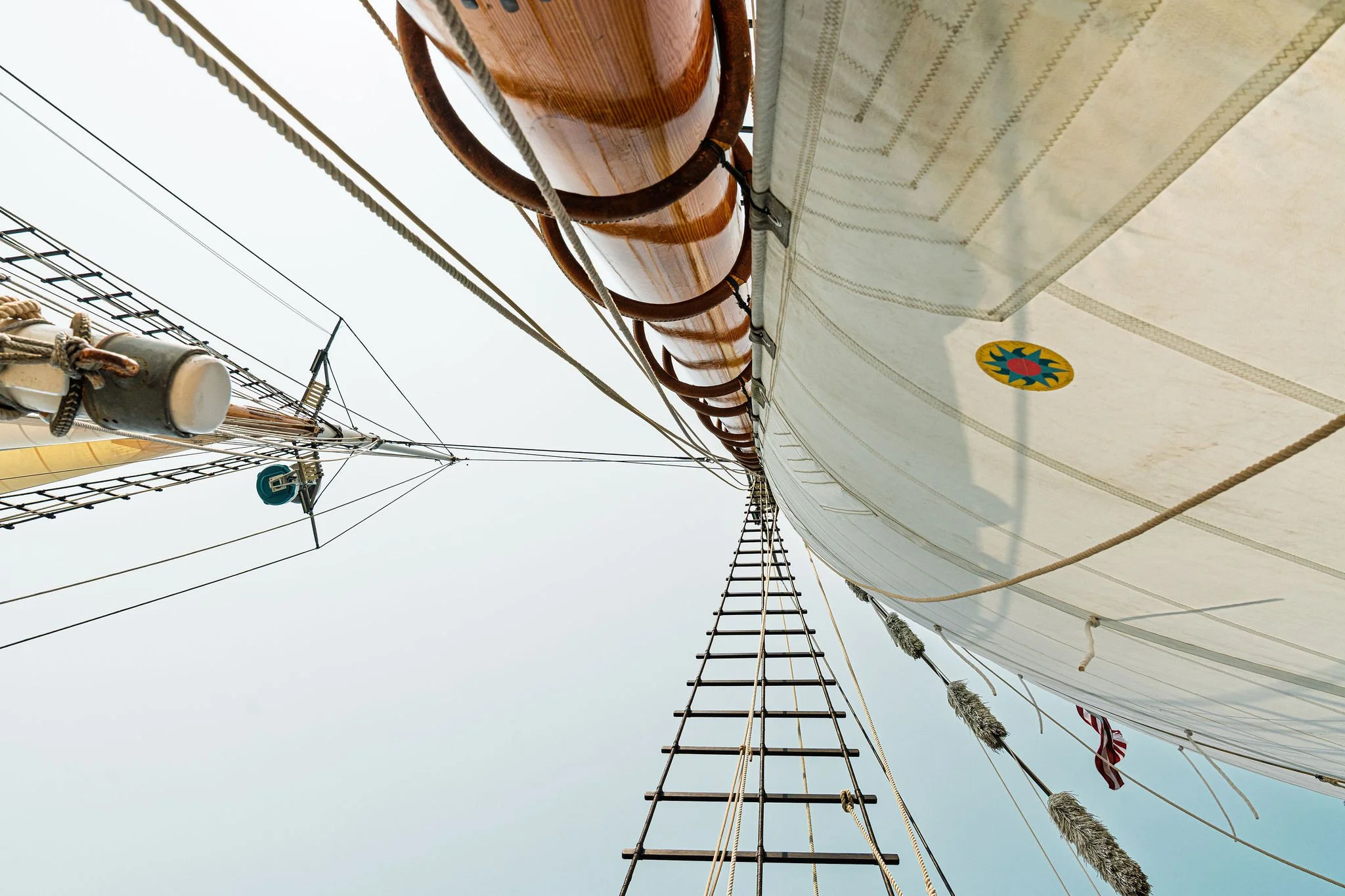 Looking up from the deck of a sailing ship at the mast, rigging, and sails against a clear sky.