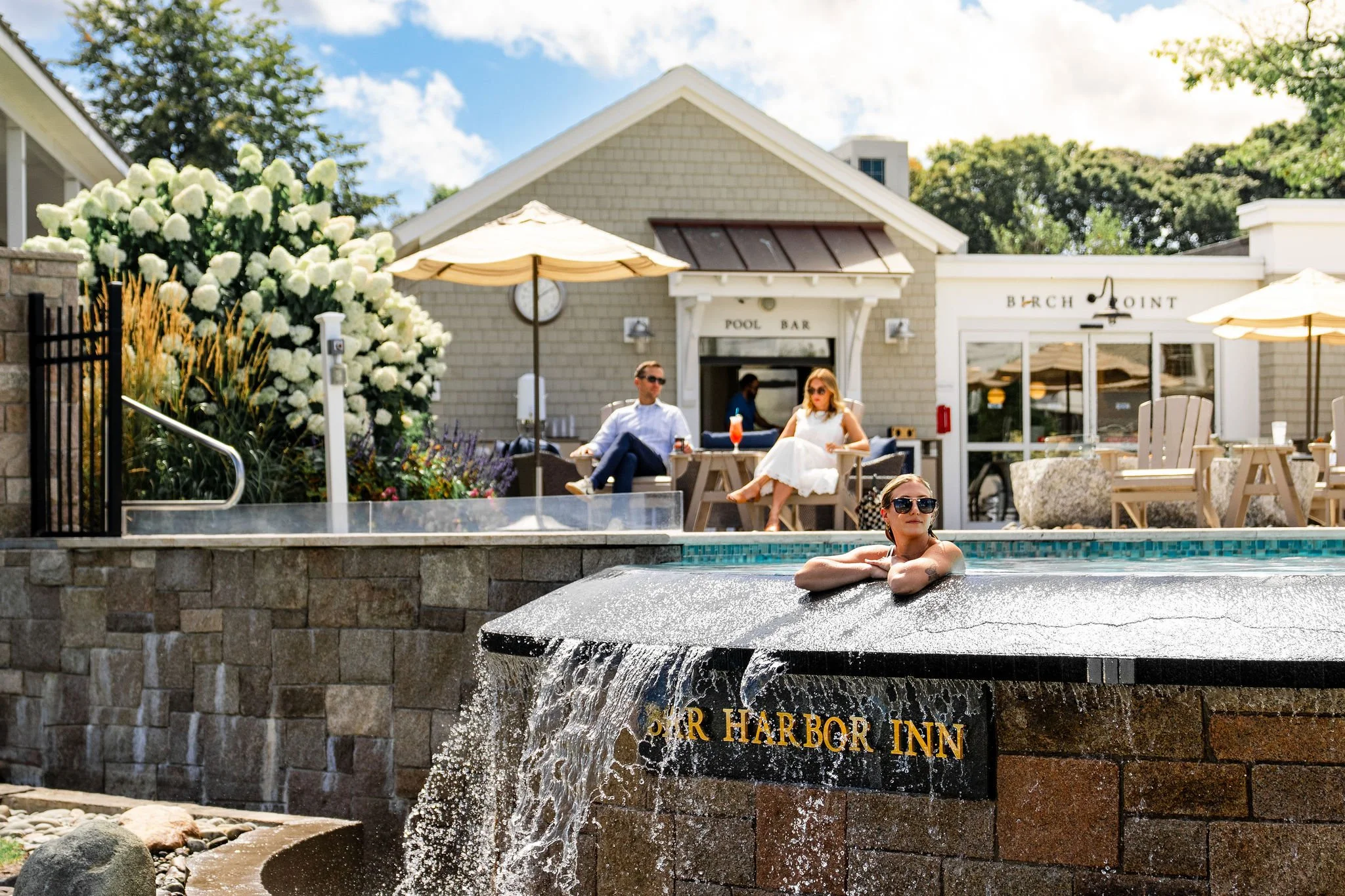 People relaxing by a pool at a resort with a sign that reads 'Star Harbor Inn'. One woman in sunglasses is in the pool, while two people sit at a table nearby, enjoying drinks on a sunny day.