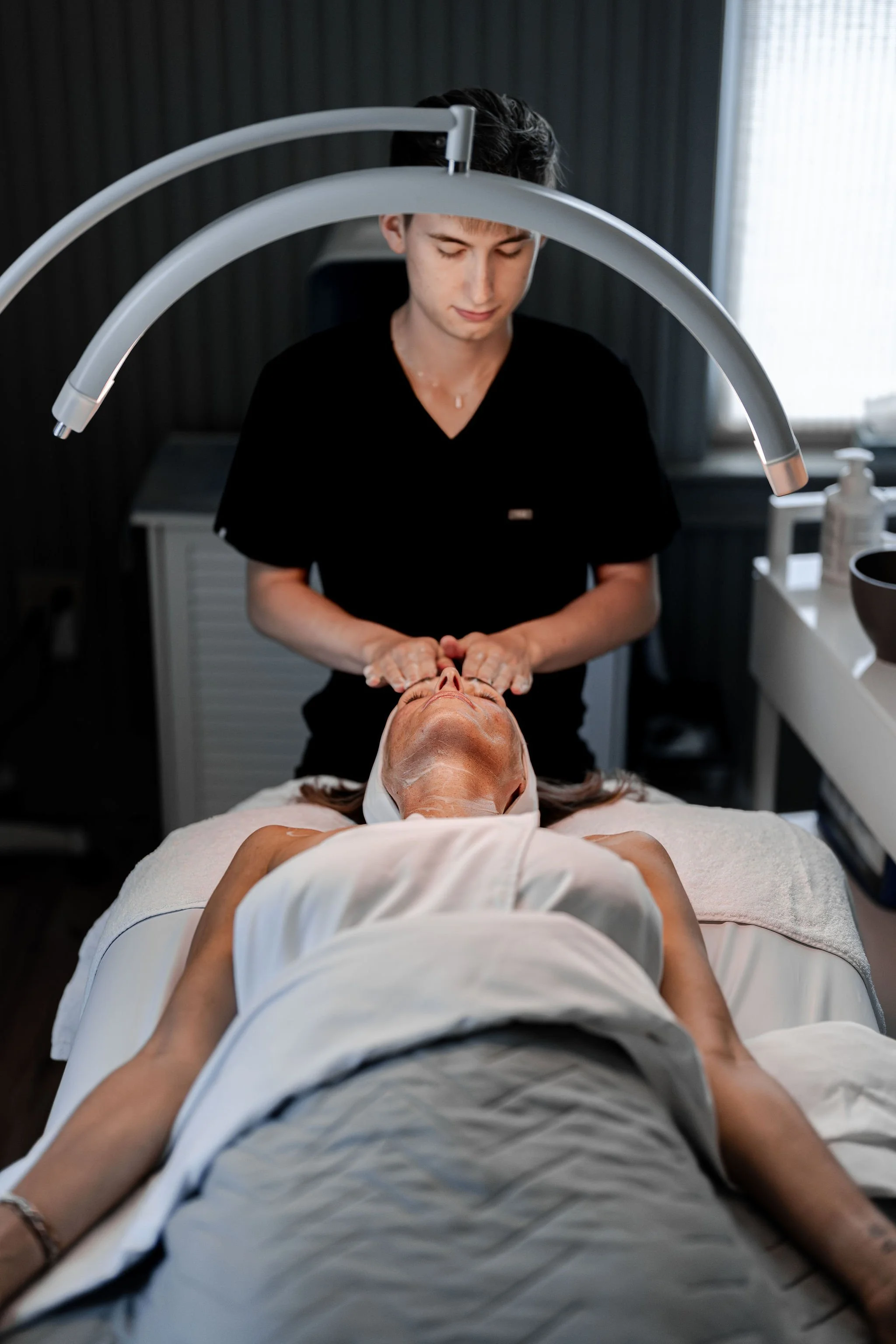 A woman lying on a spa bed receiving a facial treatment from a skincare professional in a dimly lit treatment room.