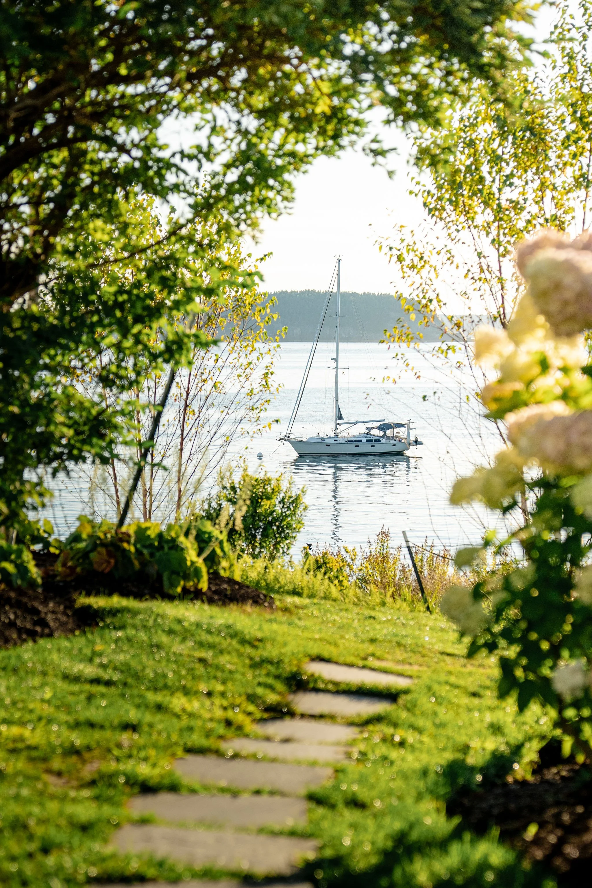 A peaceful waterfront scene with a sailboat anchored in calm water, viewed through lush greenery and flowering bushes, with a stone pathway leading towards the water.