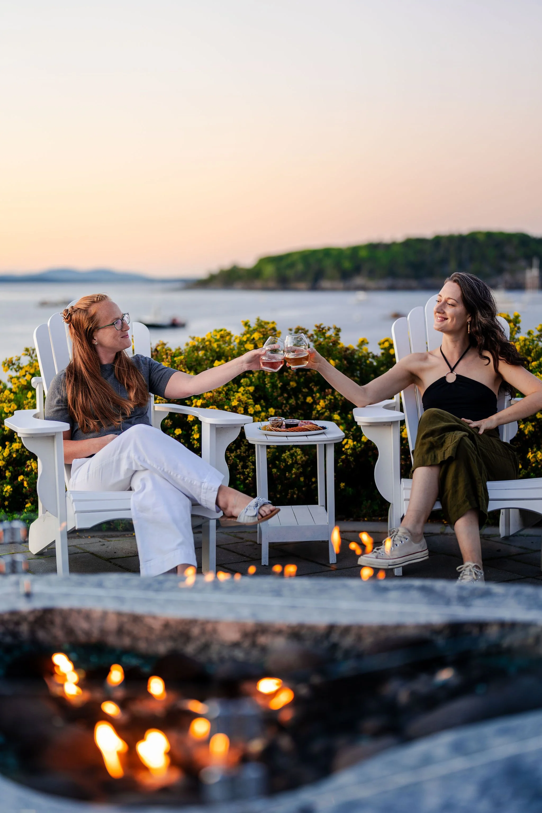 Two women are sitting outdoors on white Adirondack chairs, clinking glasses of wine or beer, at sunset near a body of water with boats, with a firepit in the foreground and yellow flowering bushes in the background.
