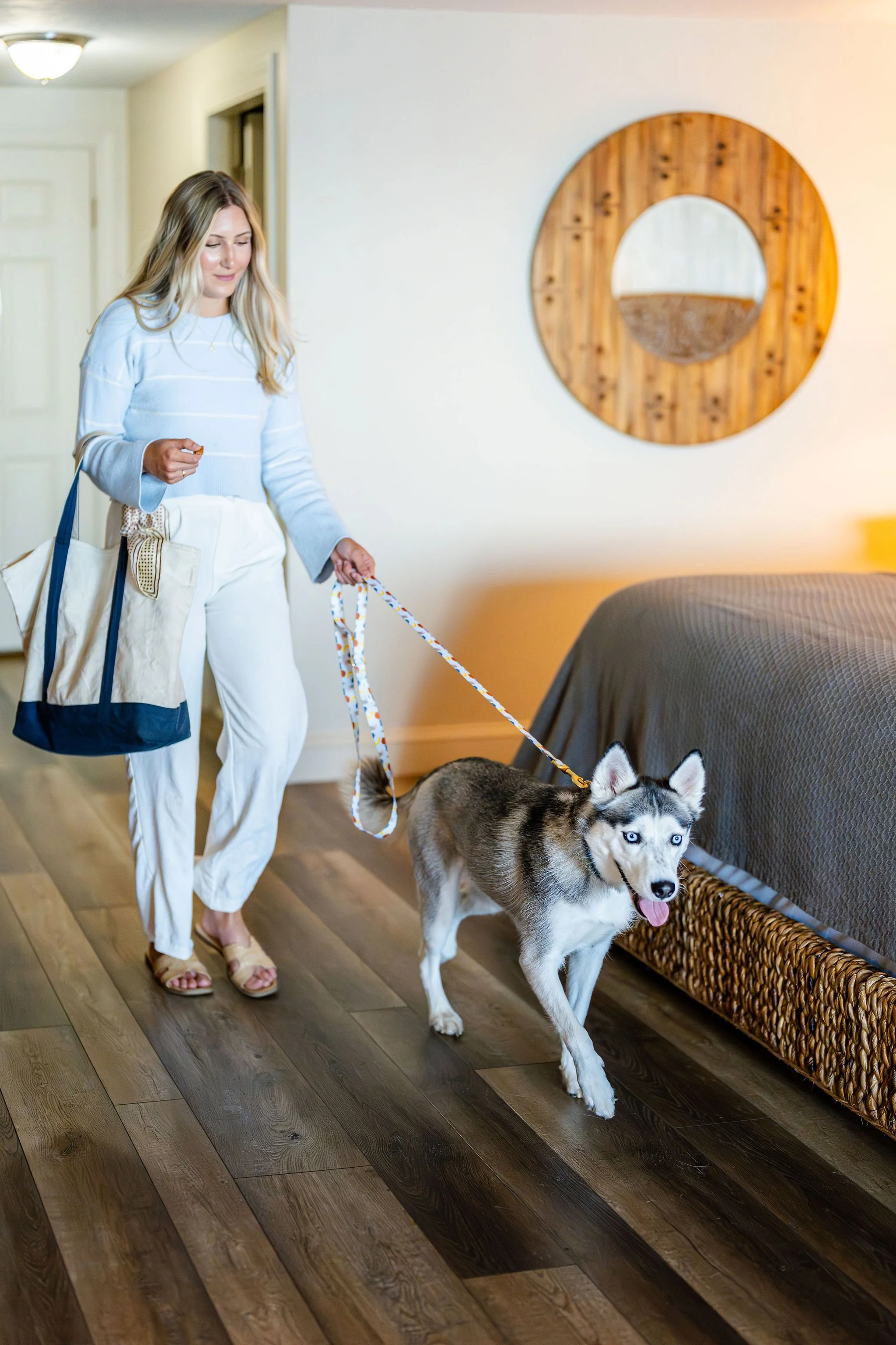 A woman walking a husky dog on a leash inside a bedroom with wooden and grey decor.