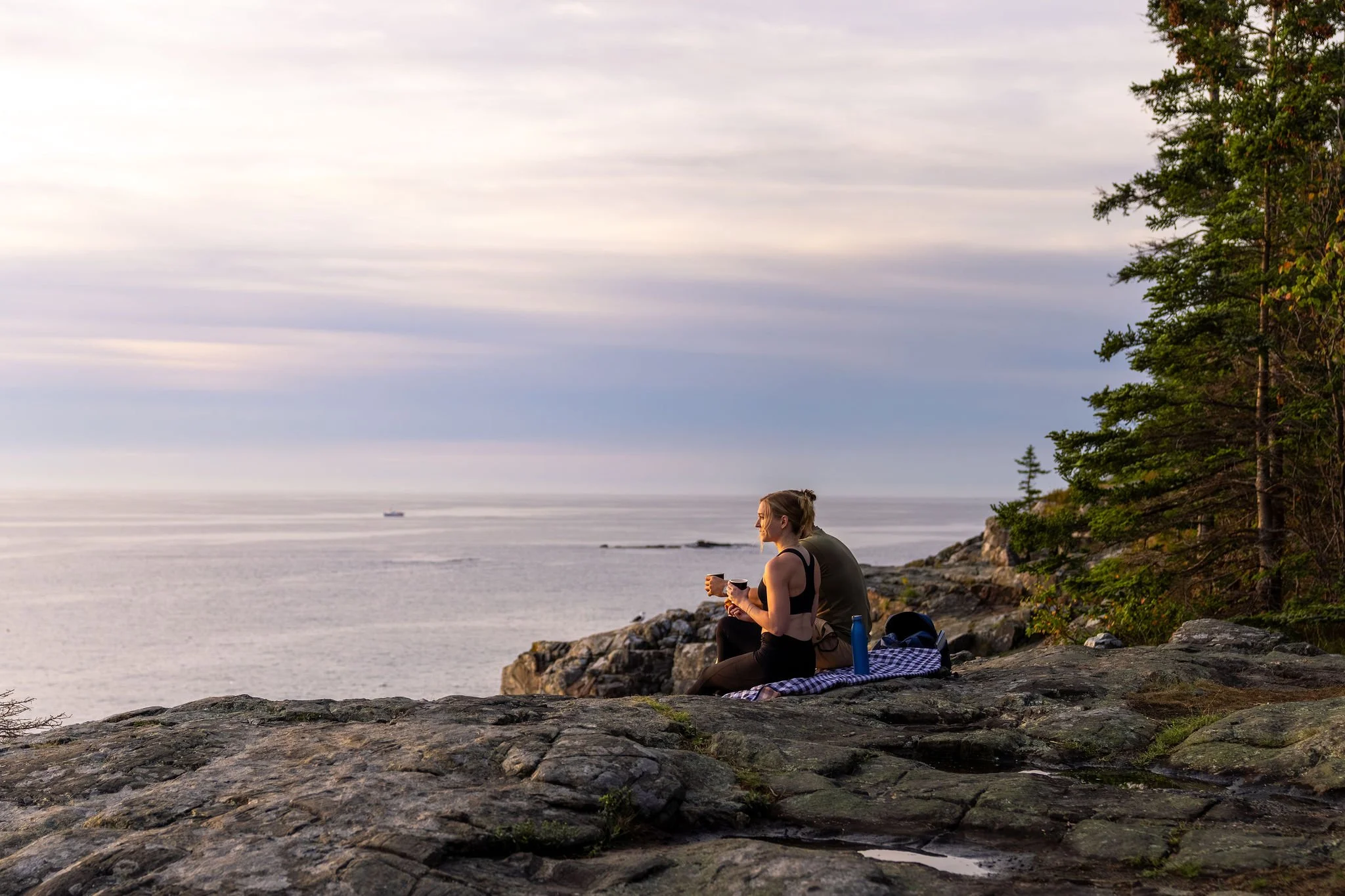 A woman and man sitting on rocks at a coastal cliff, enjoying the sunset with a woman drinking from a mug, surrounded by trees.