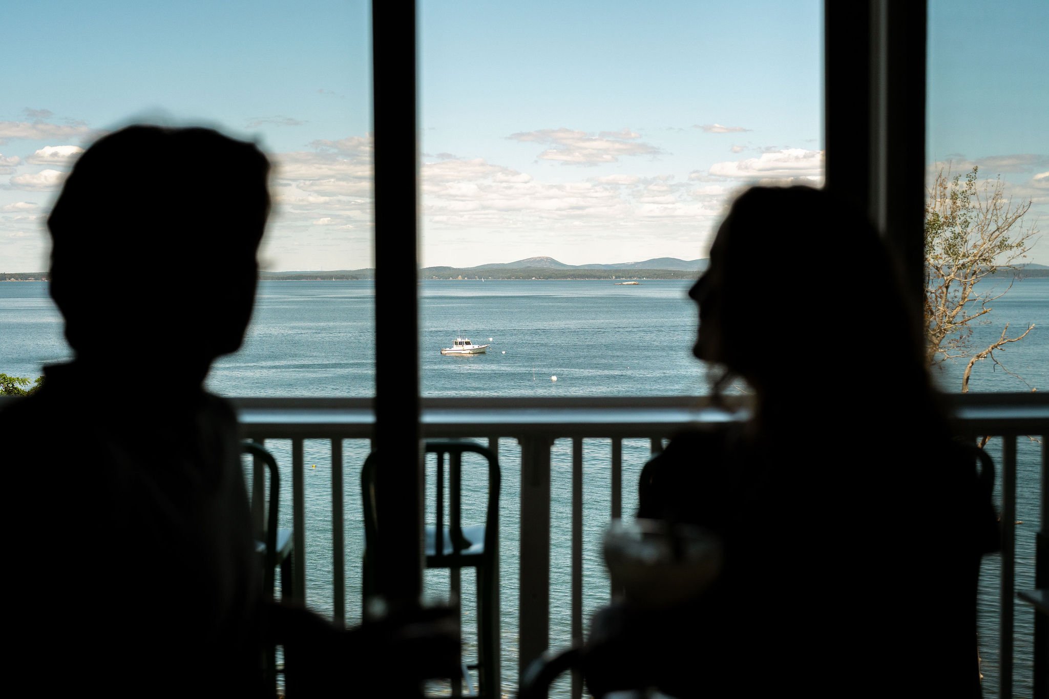 Silhouettes of a man and woman sitting at a table near a window with a view of a lake or sea, a boat in the water, and mountains in the distance.