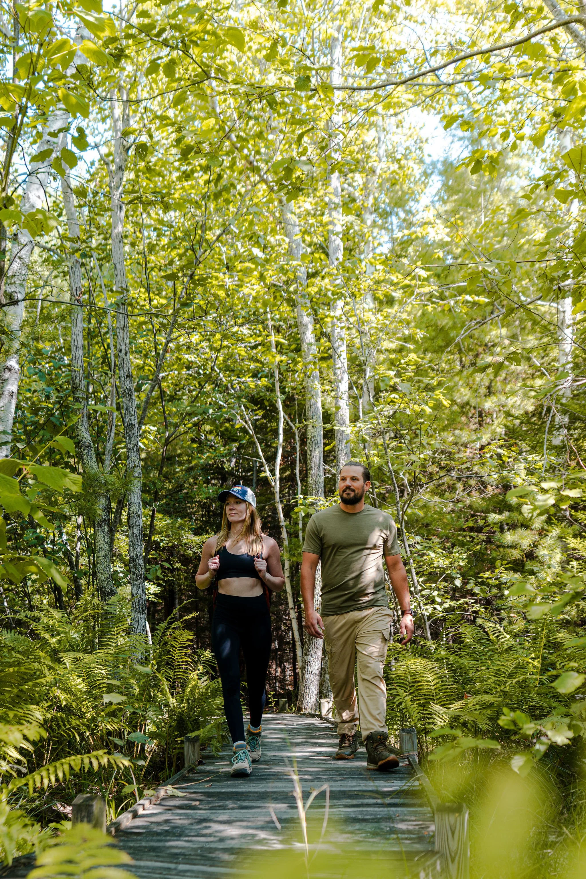 A man and a woman hiking on a wooden trail through a lush green forest with tall trees and dense foliage.