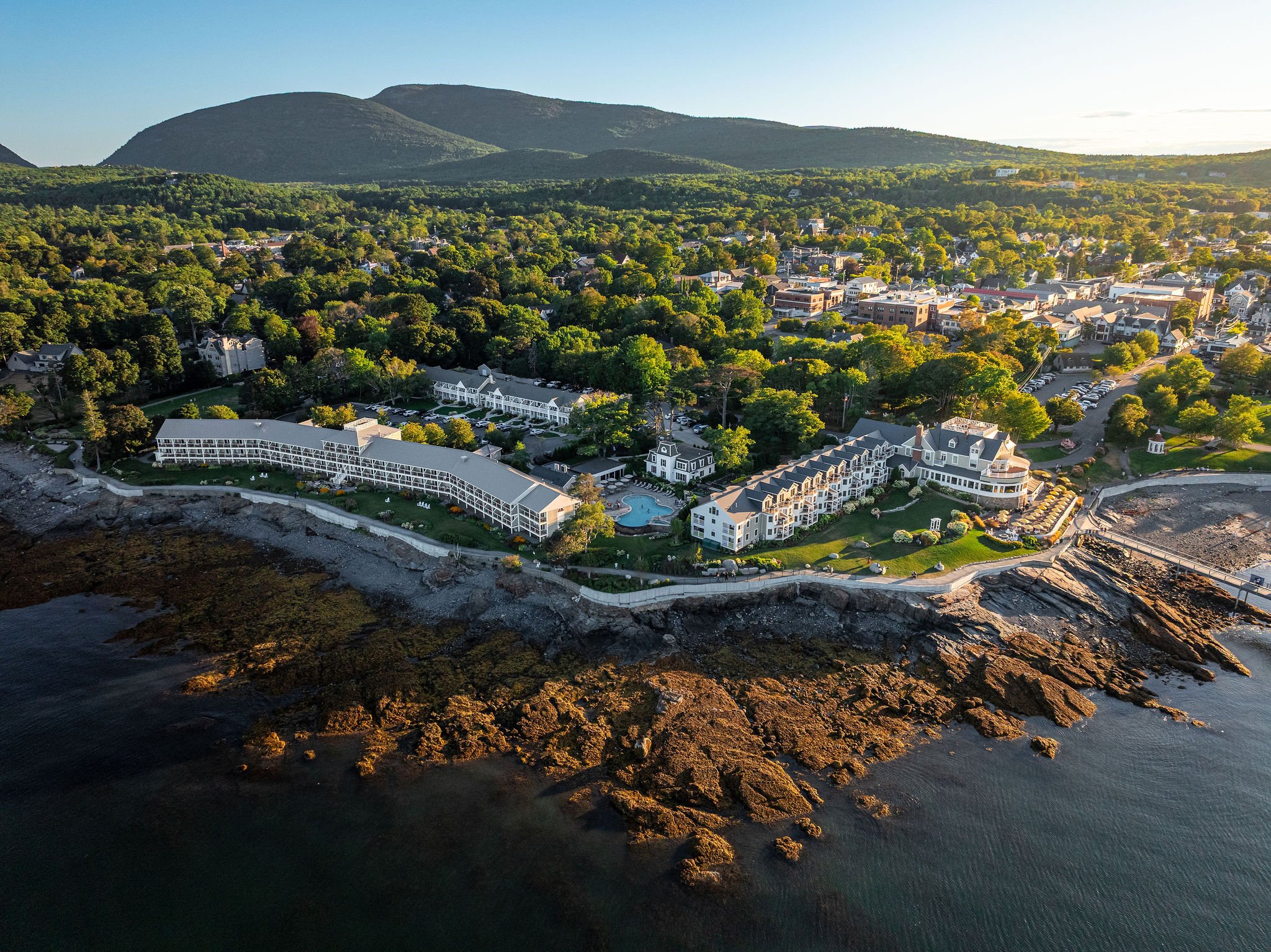 Aerial view of a coastal town with residential buildings, a pool, and lush green trees, with mountains in the background and water in the foreground.