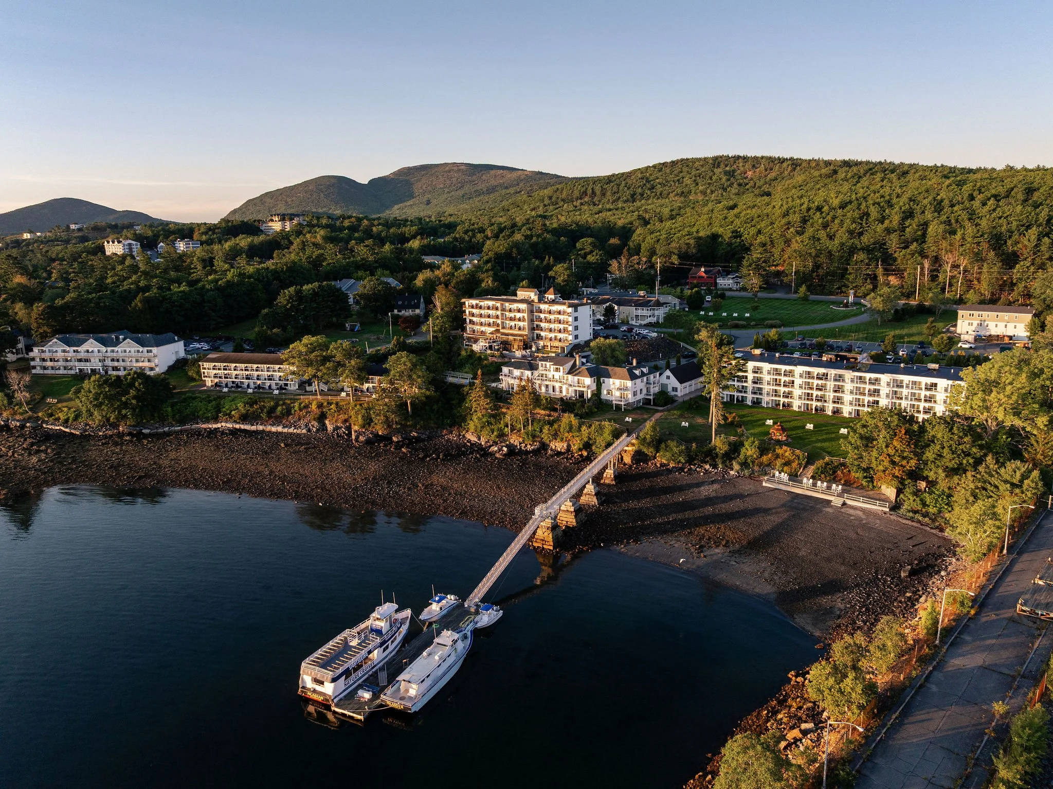 Aerial view of a coastal resort with multiple white and beige buildings, a dock with two boats, a rocky shoreline, green hills, and forested mountains in the background during sunset.