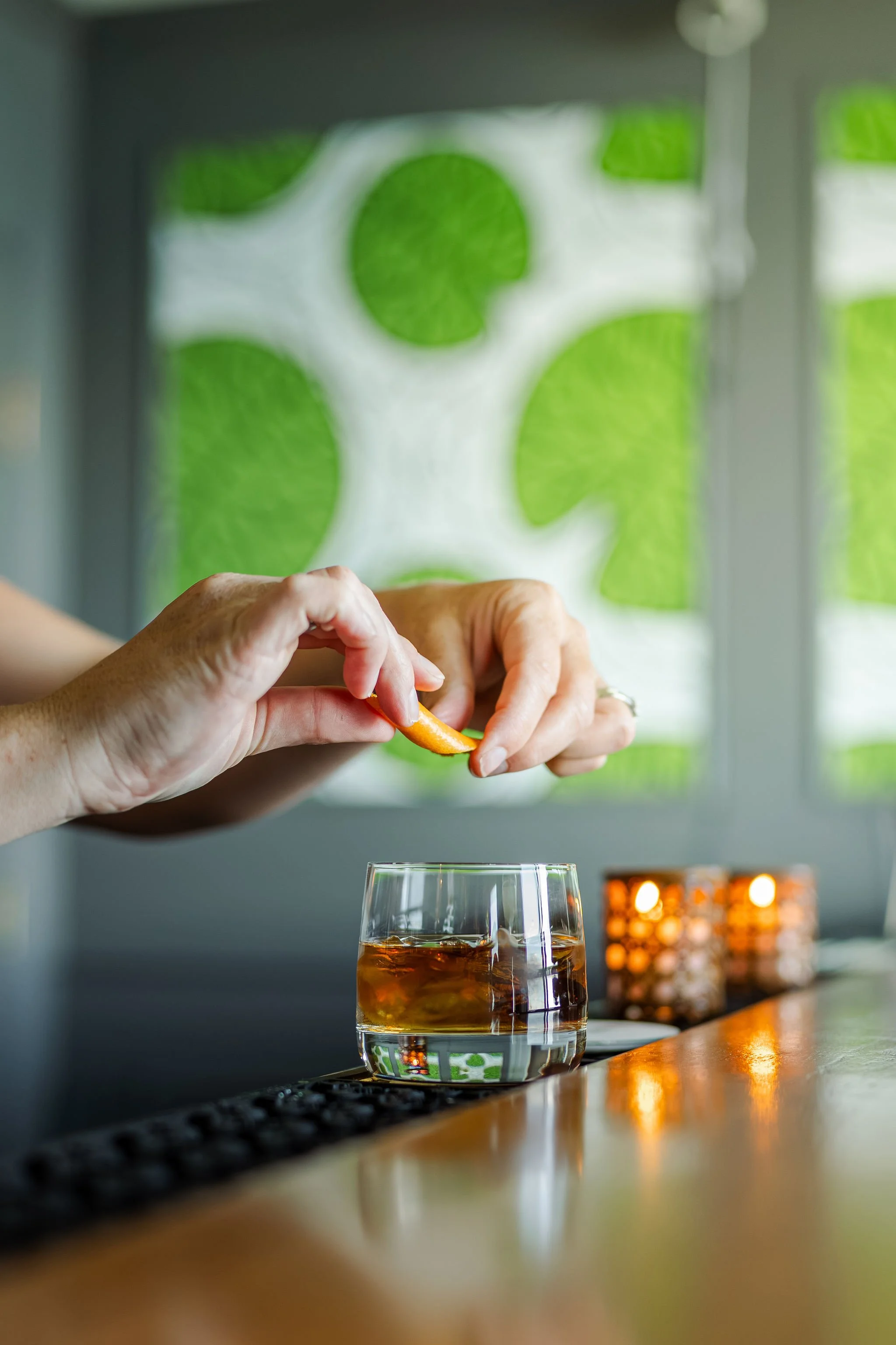 A person squeezing a citrus fruit peel into a glass of whiskey on a bar counter, with candles and a green and white abstract wall in the background.