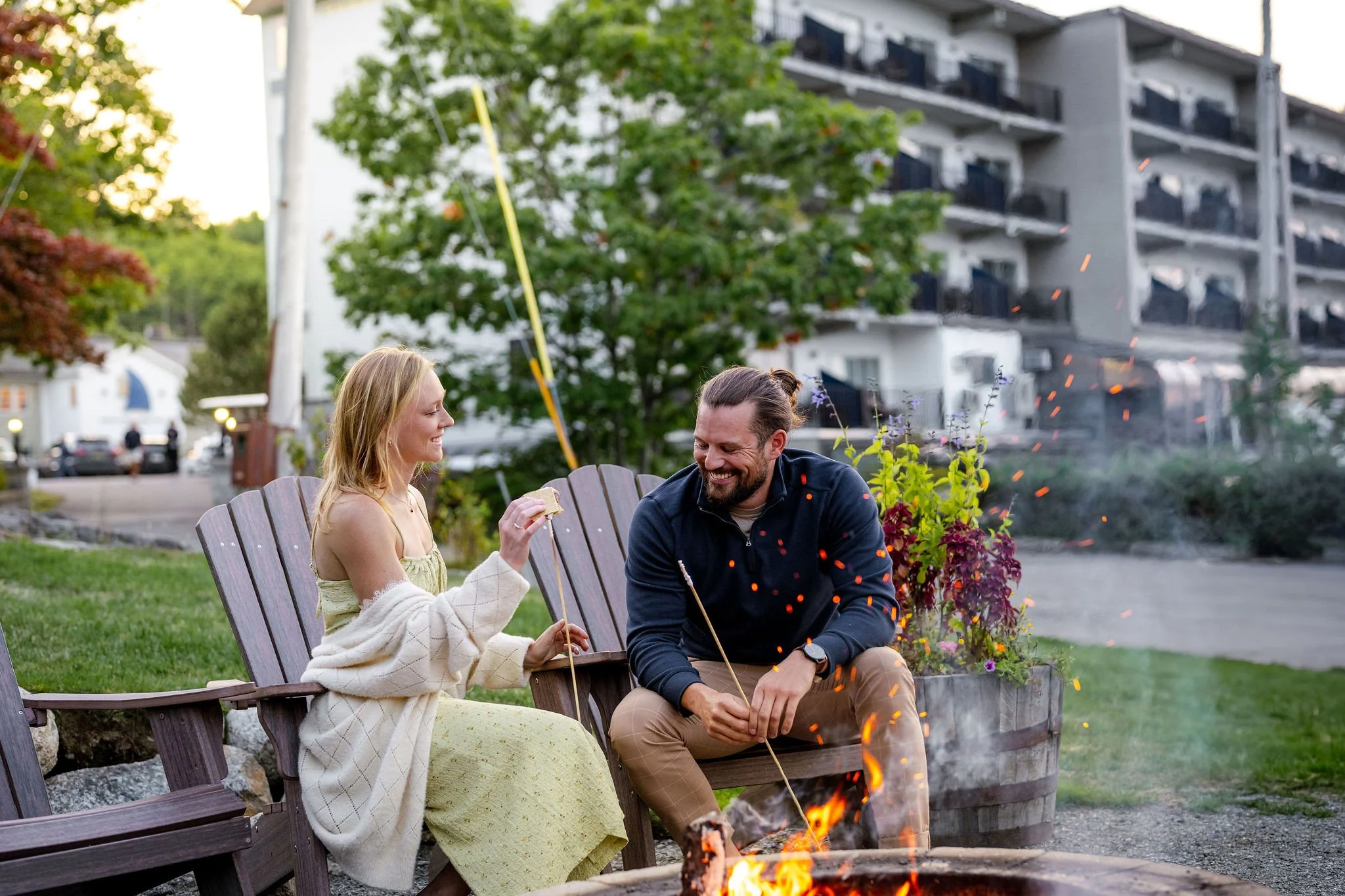 A man and woman sitting on wooden chairs around a campfire outdoors, smiling and roasting marshmallows, with a multi-story building and trees in the background.