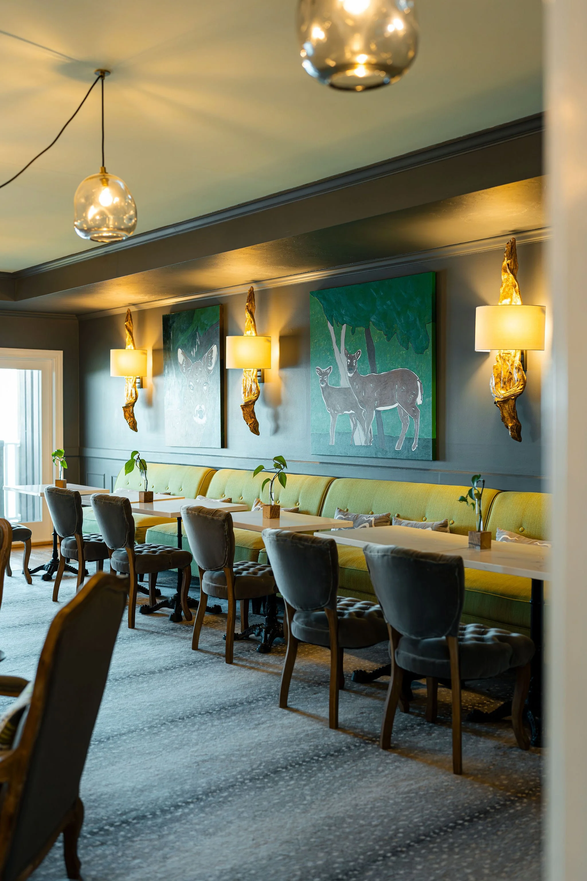 Interior of a restaurant with a long yellow cushioned bench and several gray velvet chairs, decorated with small potted plants on white tables, wall art of deer, and warm lighting fixtures.