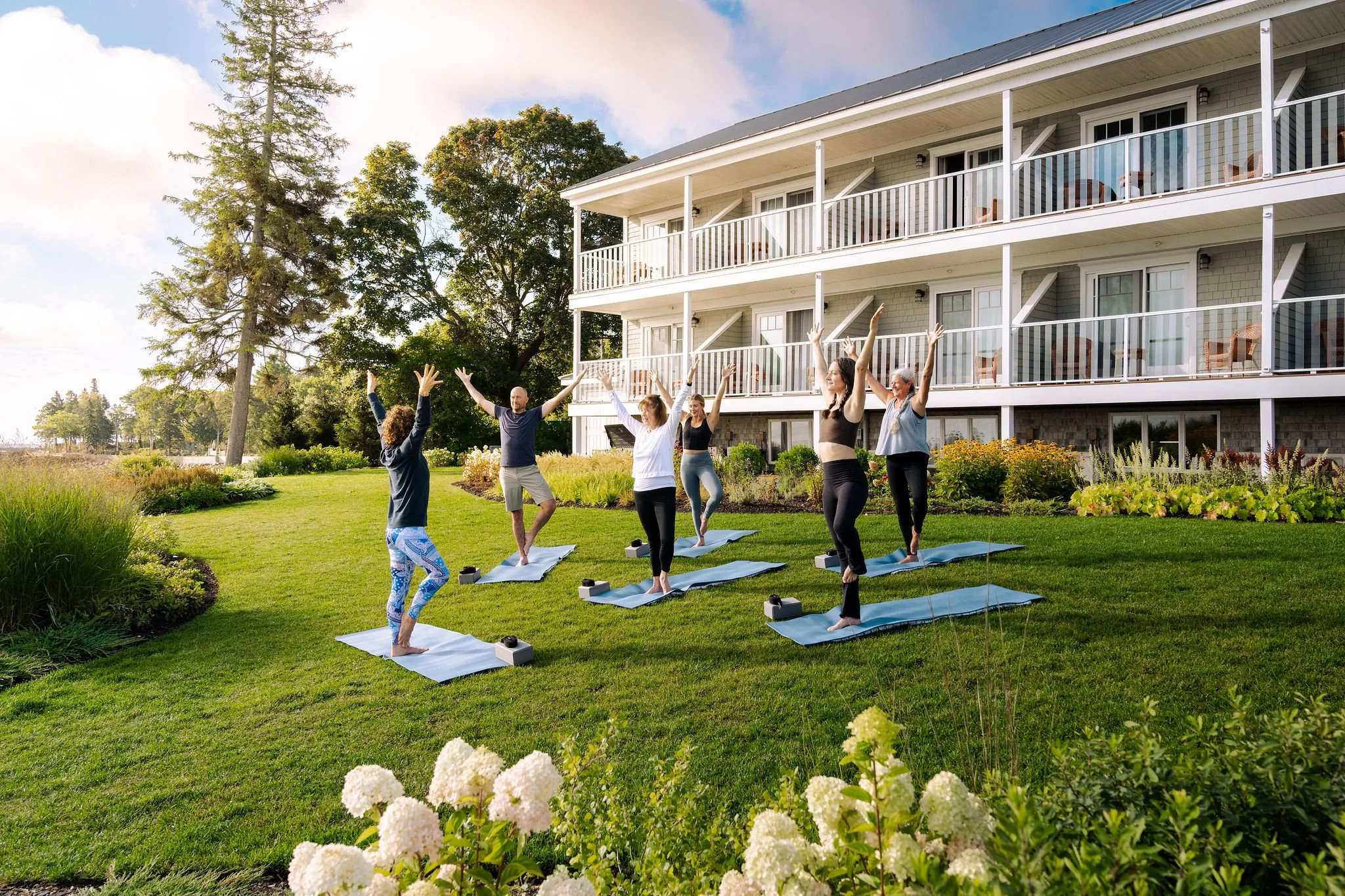 Group of six people practicing yoga outdoors on a grassy lawn in front of a multi-story building with balconies, during daytime with partly cloudy sky.
