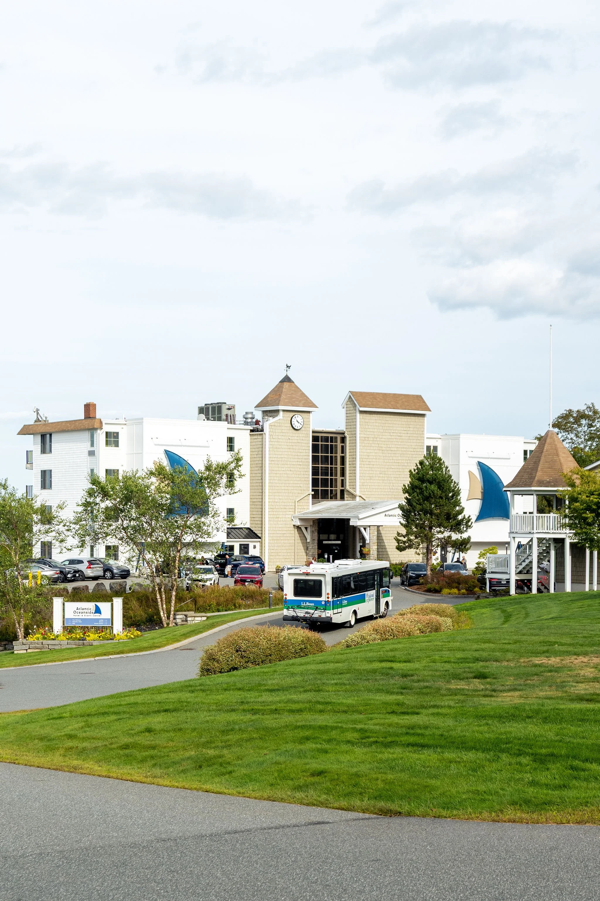 A white bus driving on a curved road towards a building with a clock tower, surrounded by green lawns, trees, and parked cars, with a partly cloudy sky above.