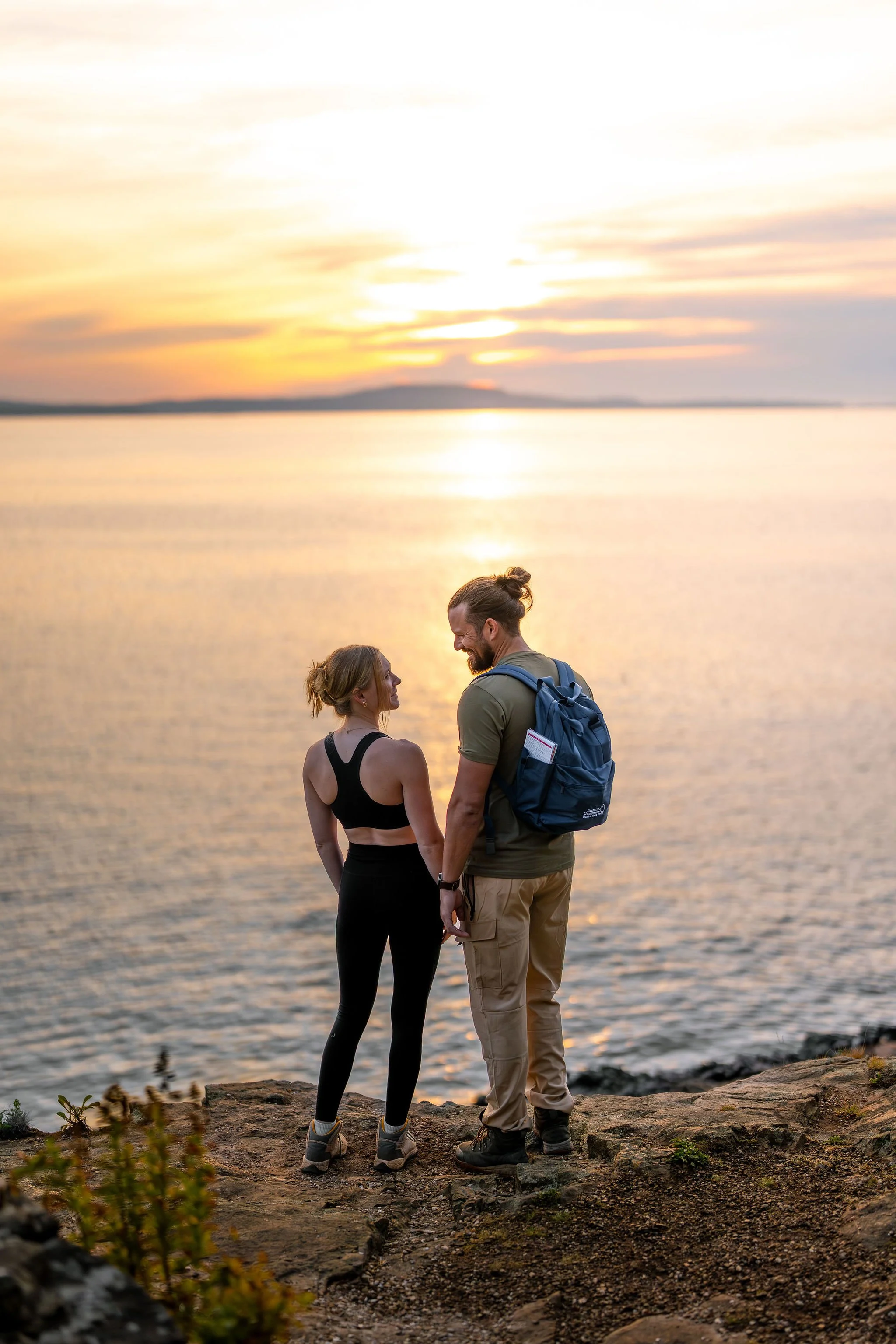 A couple standing on a rocky shoreline at sunset, holding hands and looking at each other by a calm body of water.