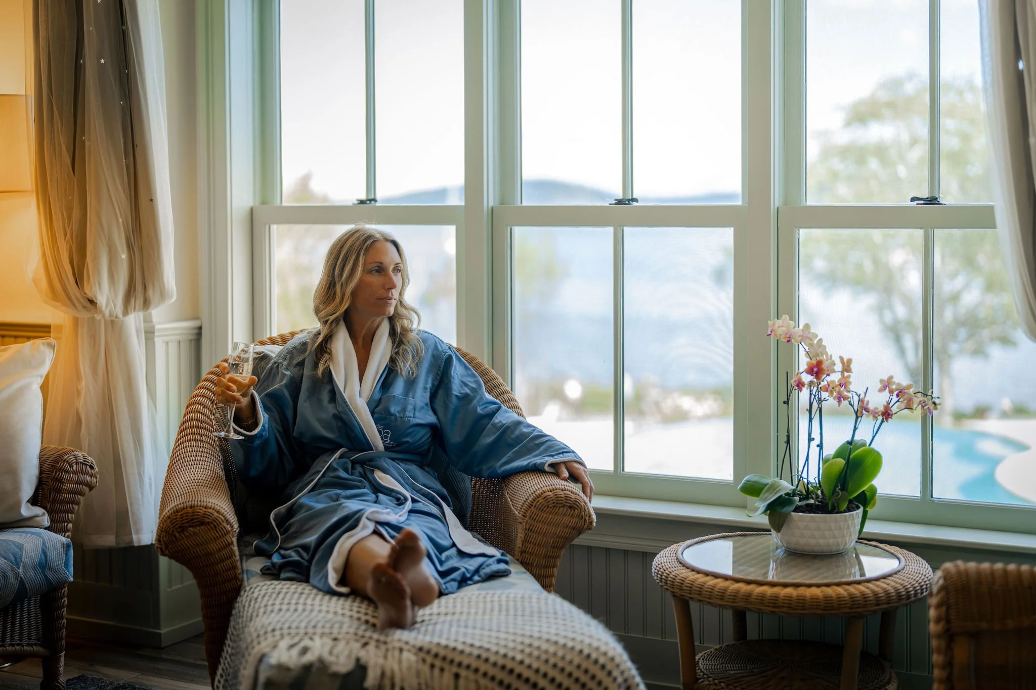 A woman in a blue bathrobe sitting on a wicker chair, holding a glass of wine, near a large window with curtains, in a cozy indoor setting with a plant on a round table.