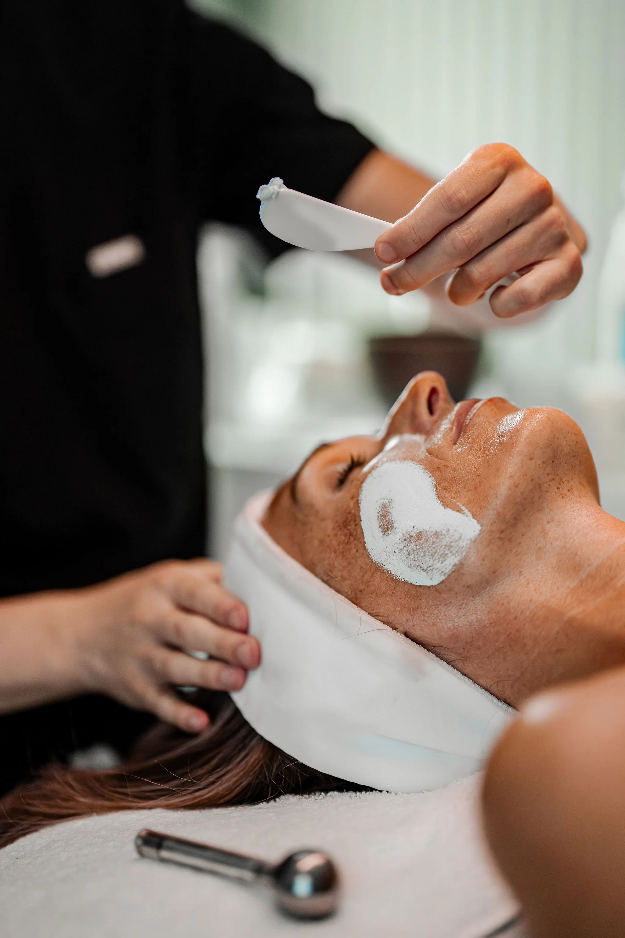 A woman receiving a facial treatment with a white facial mask applied; a cosmetologist holds a spatula or tool over her face.