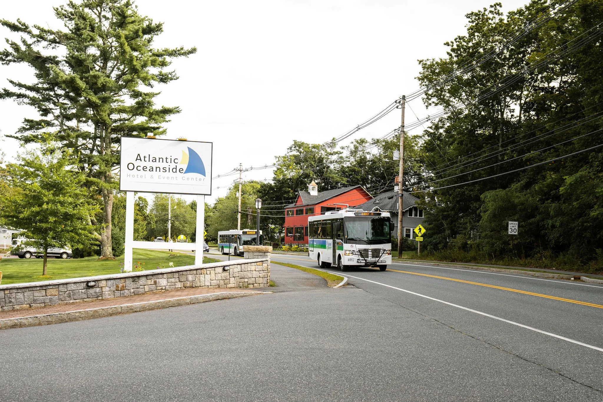 Street view with a bus in front of a sign for Atlantic Oceanside Hotel & Event Center, trees, houses, and utility poles on a cloudy day.