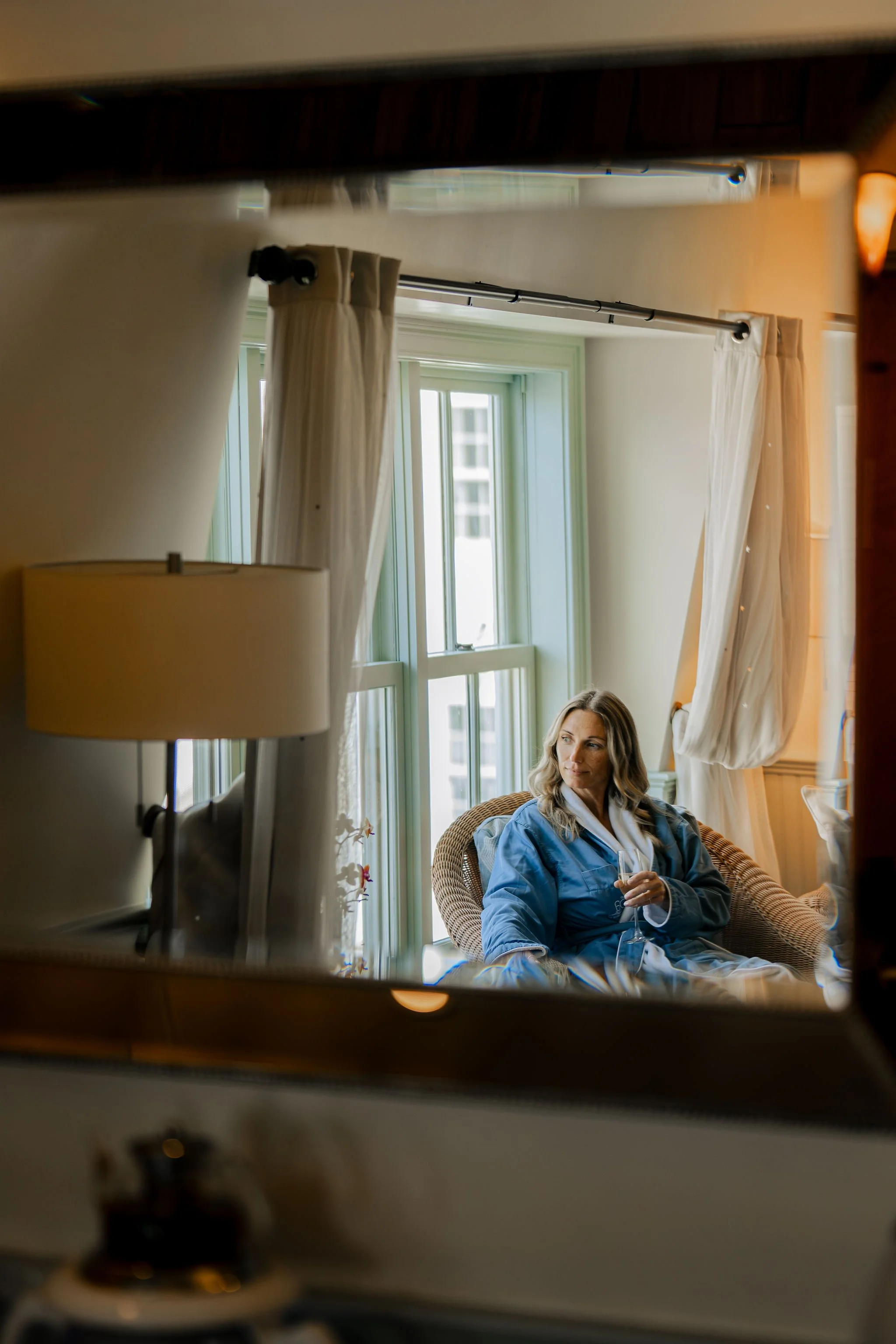 Woman sitting by a window with cream curtains, holding a glass of wine, seen through a mirror.