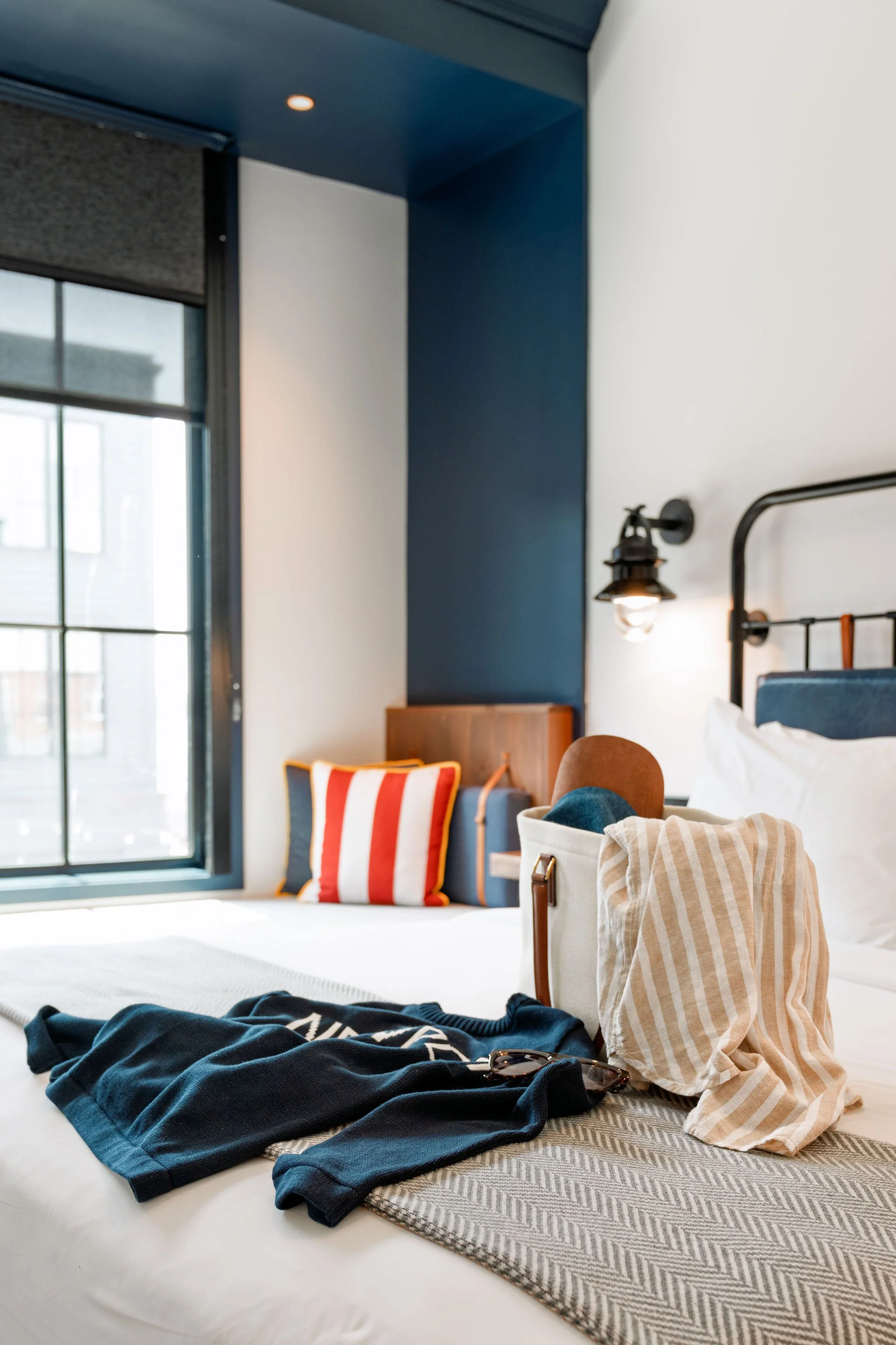 Interior of a bedroom with a window, a bed with a beige striped towel, dark sunglasses, a bag, a sweater, and colorful pillows. Modern decor with a blue and white color scheme.