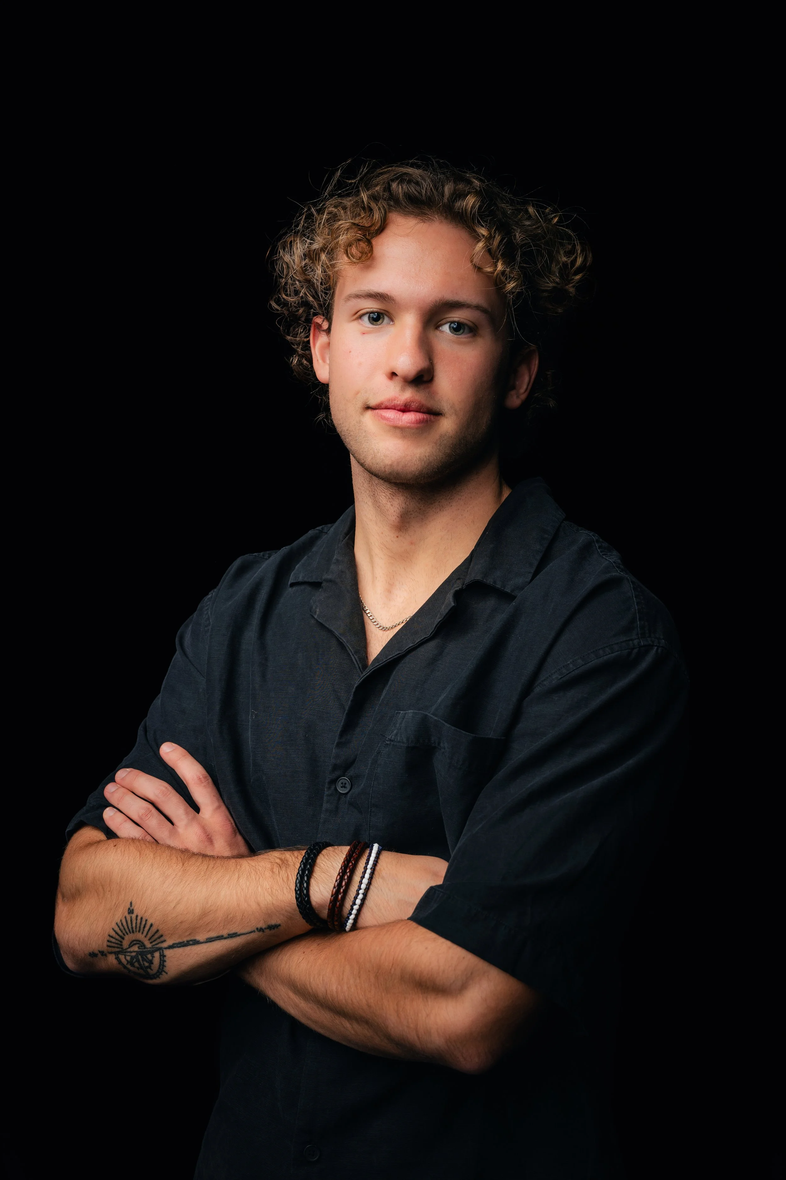 Young man with curly hair, arms crossed, wearing a black shirt and bracelets, against a black background.