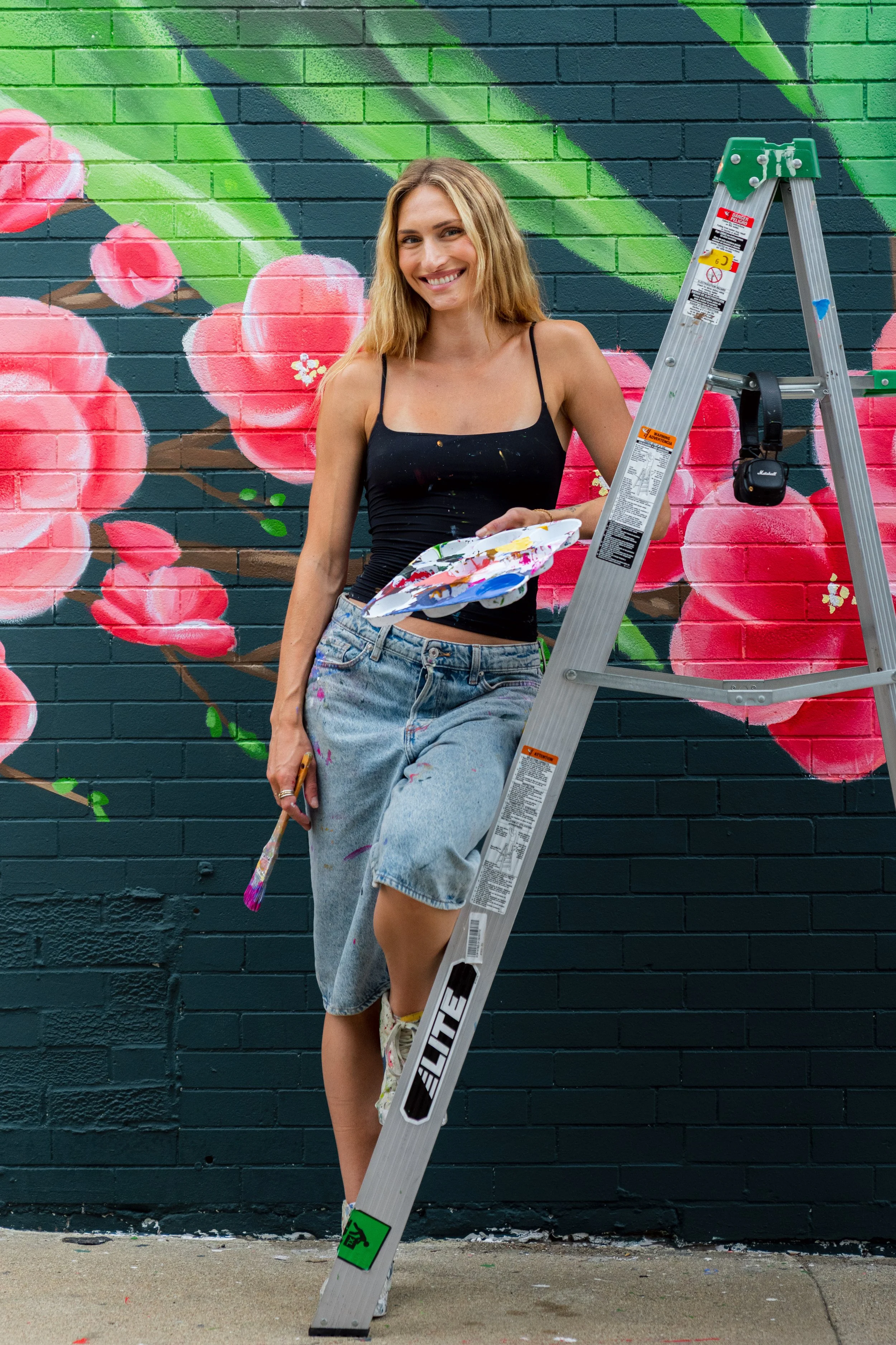 A woman standing next to a ladder, painting a mural of pink flowers and green leaves on a brick wall.