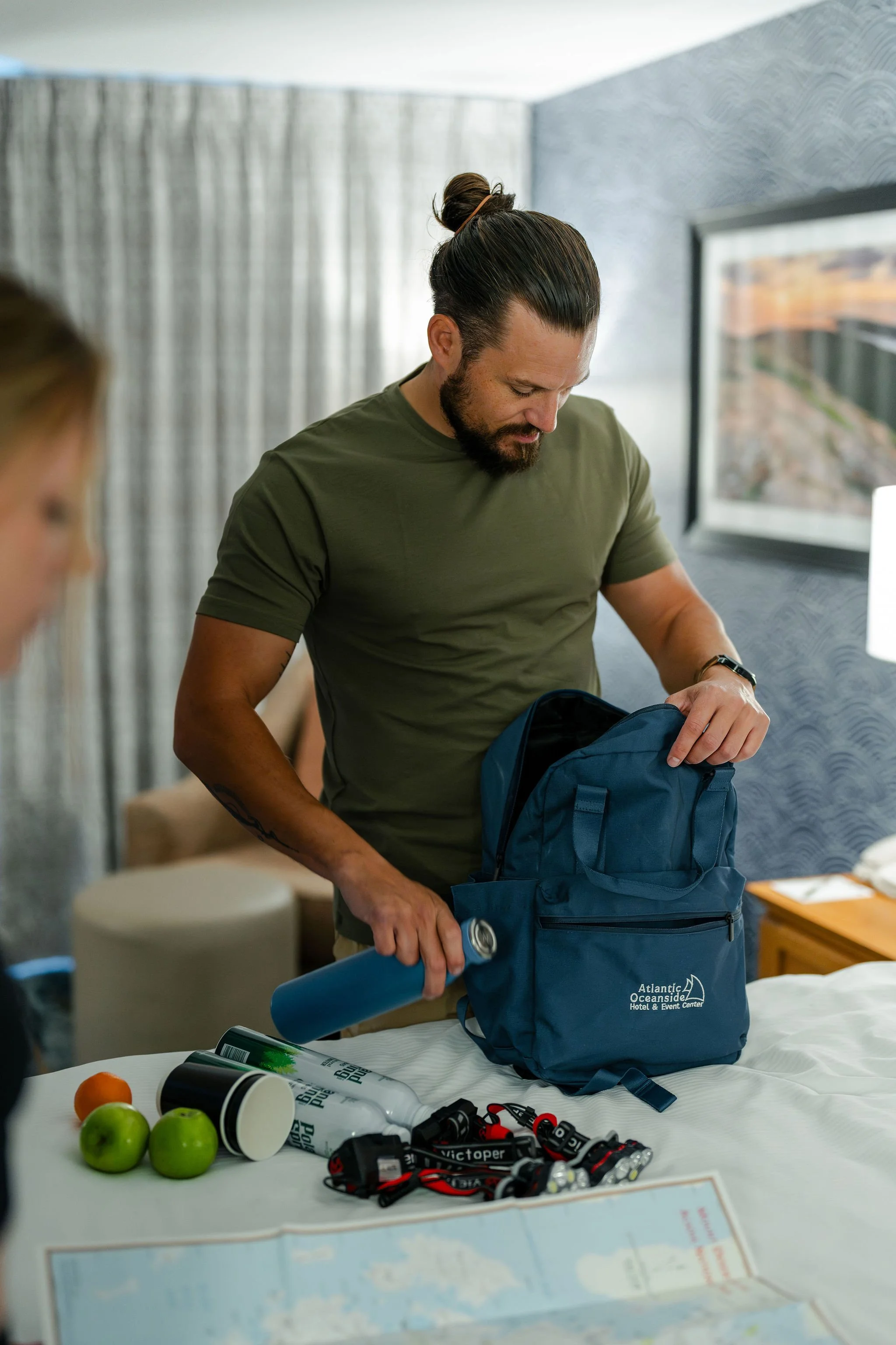 A man with a bun hairstyle and beard packs a blue backpack on a hotel bed. Items on the bed include water bottles, apples, a can, headlamps, and a map. A woman is partially visible in the foreground.
