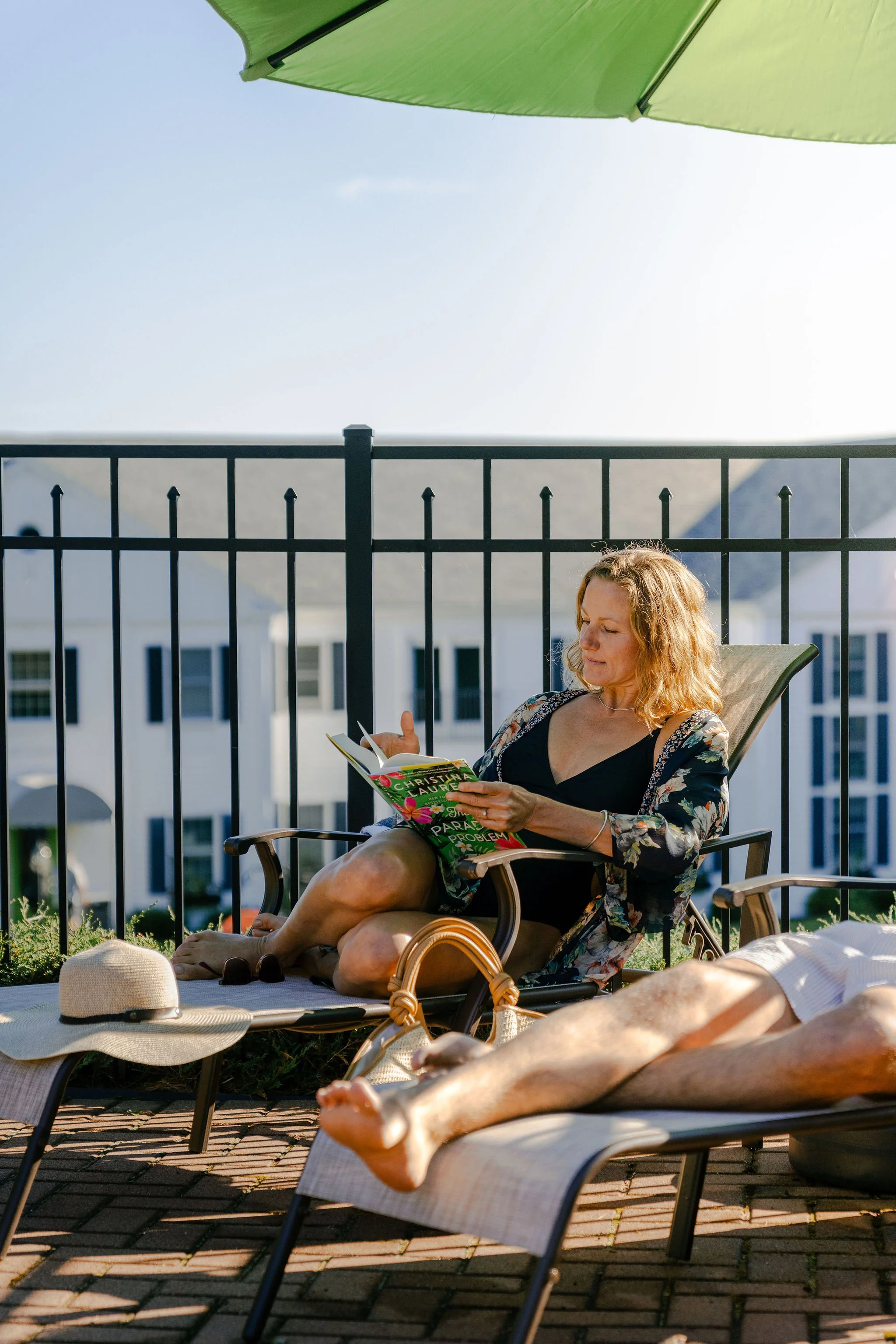A woman with blonde curly hair relaxing on a lounge chair outdoors, reading a book under a green umbrella, with another person sunbathing nearby.