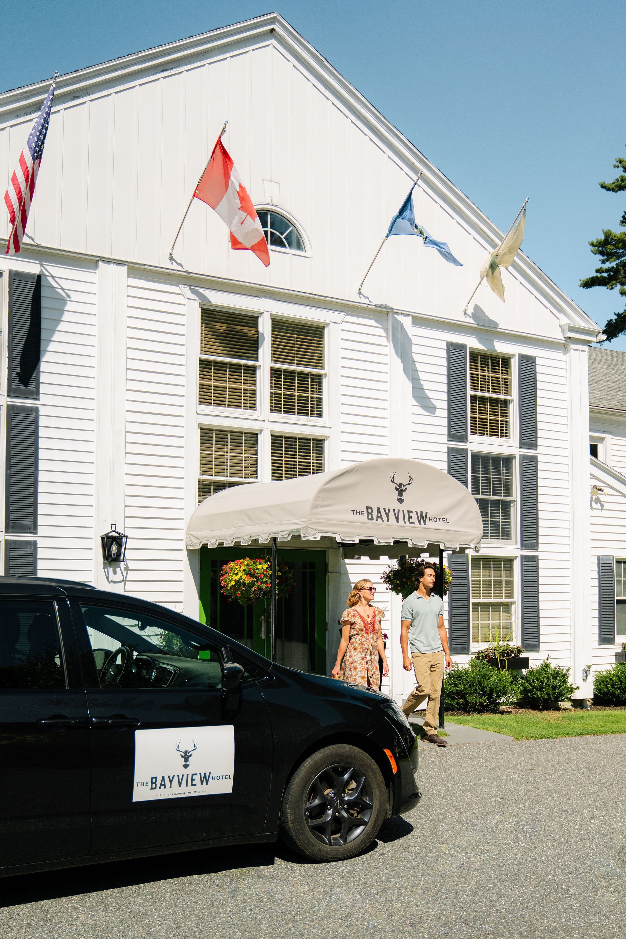 People walking out of The Bayview Hotel, a white building with flags on the front, a canopy, and a black car with the hotel logo parked in front.