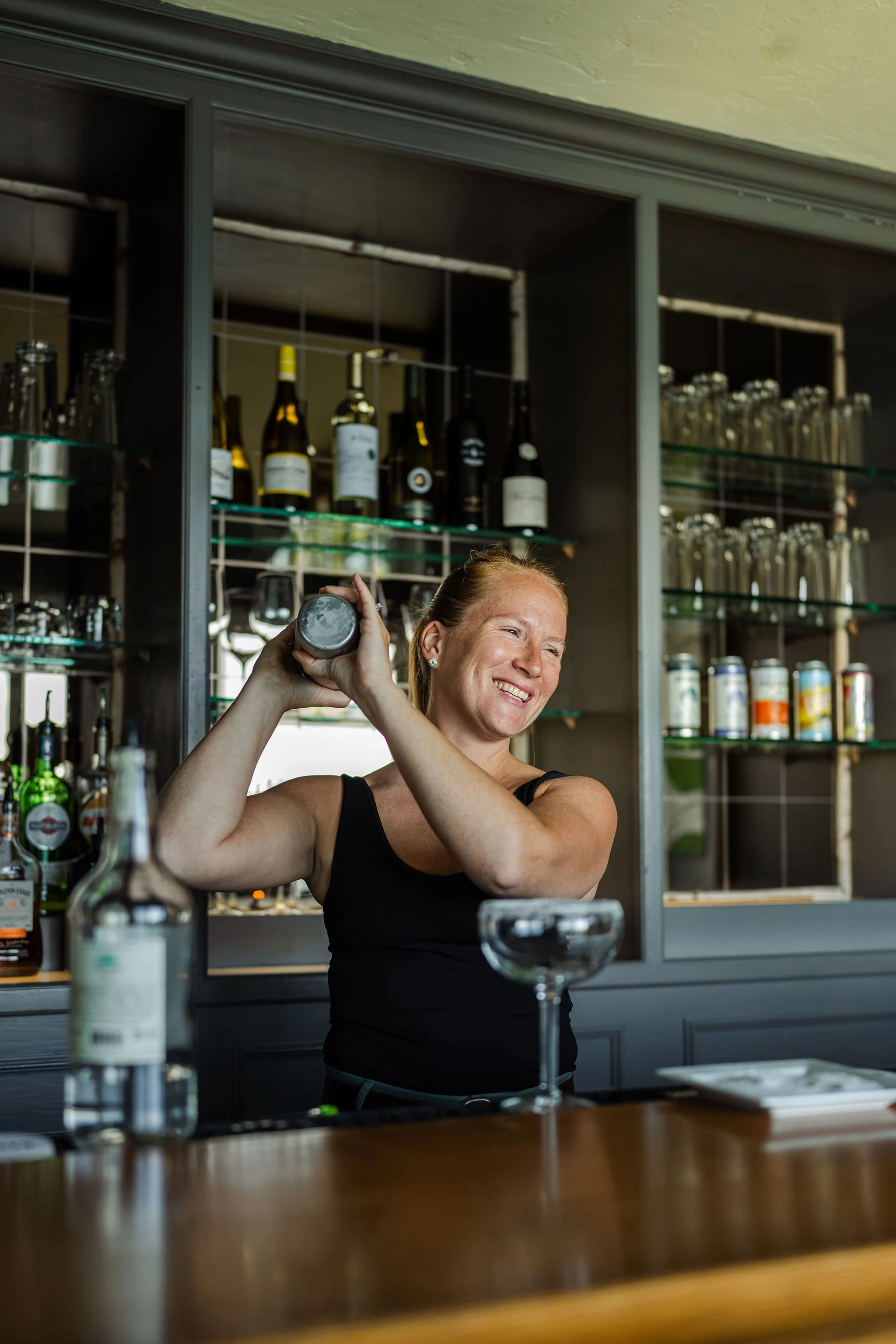 A woman behind a bar smiling as she shakes a cocktail shaker.