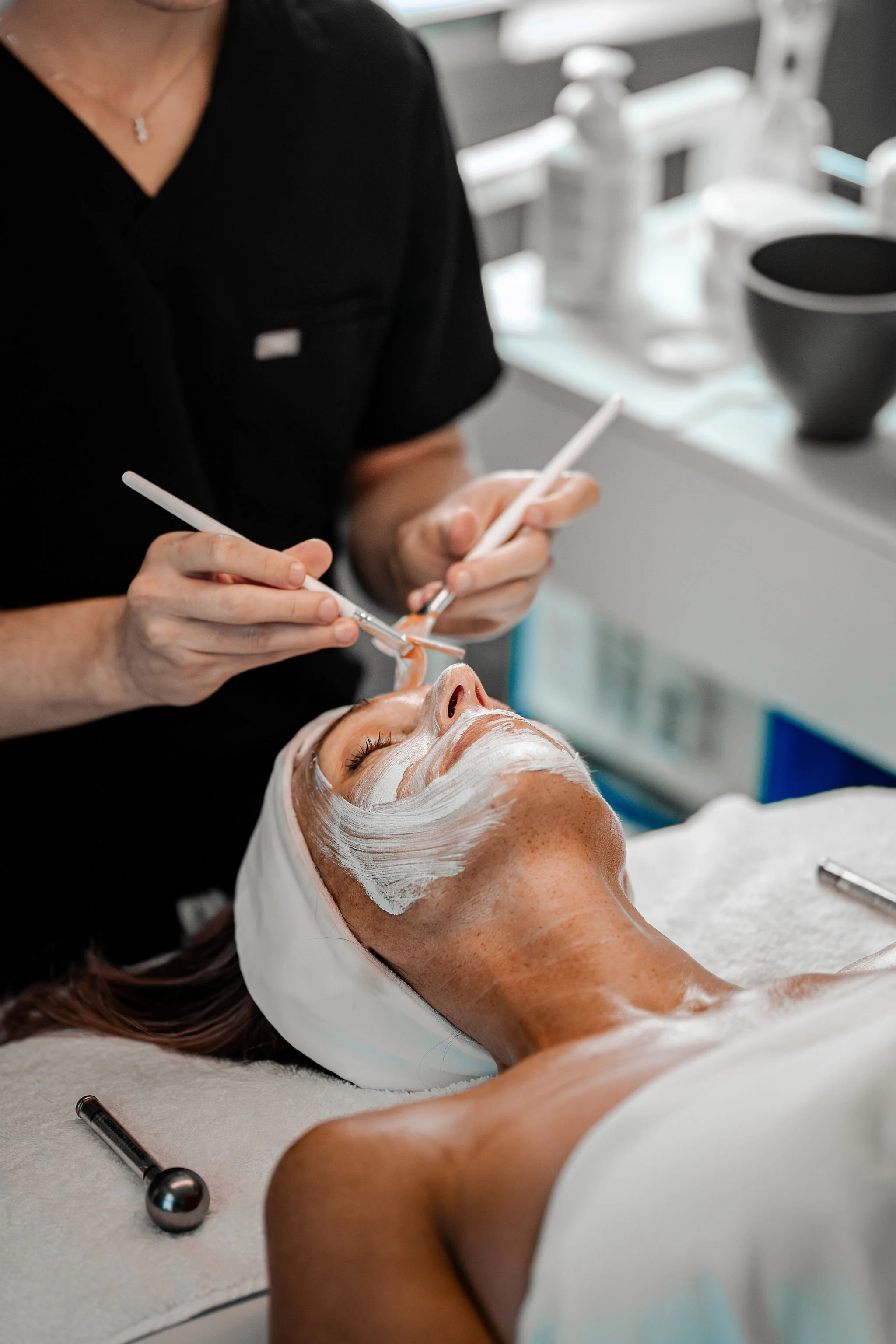 A woman receiving a facial treatment with a skincare mask applied, lying on a treatment bed while a professional applies a product to her face with tools.